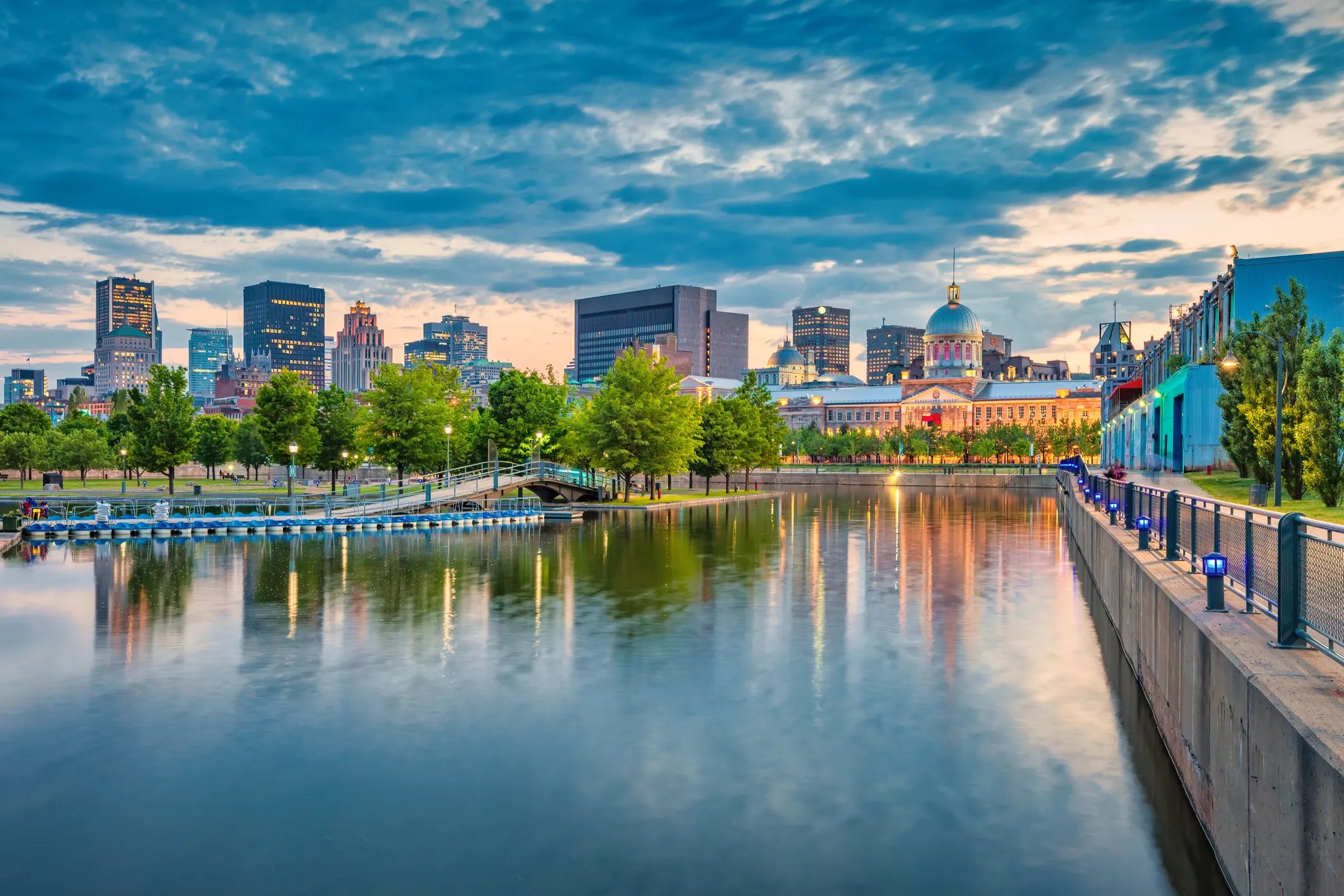 Montreal city skyline at dusk with buildings reflecting in calm waterfront near trees and a dock.