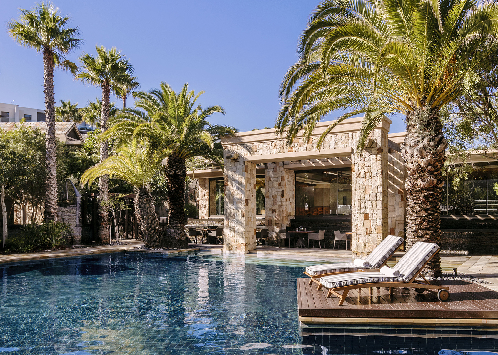 Africa, South Africa, One&Only Cape Town, sun loungers and palm trees by the pool, with poolside restaurant in the background 