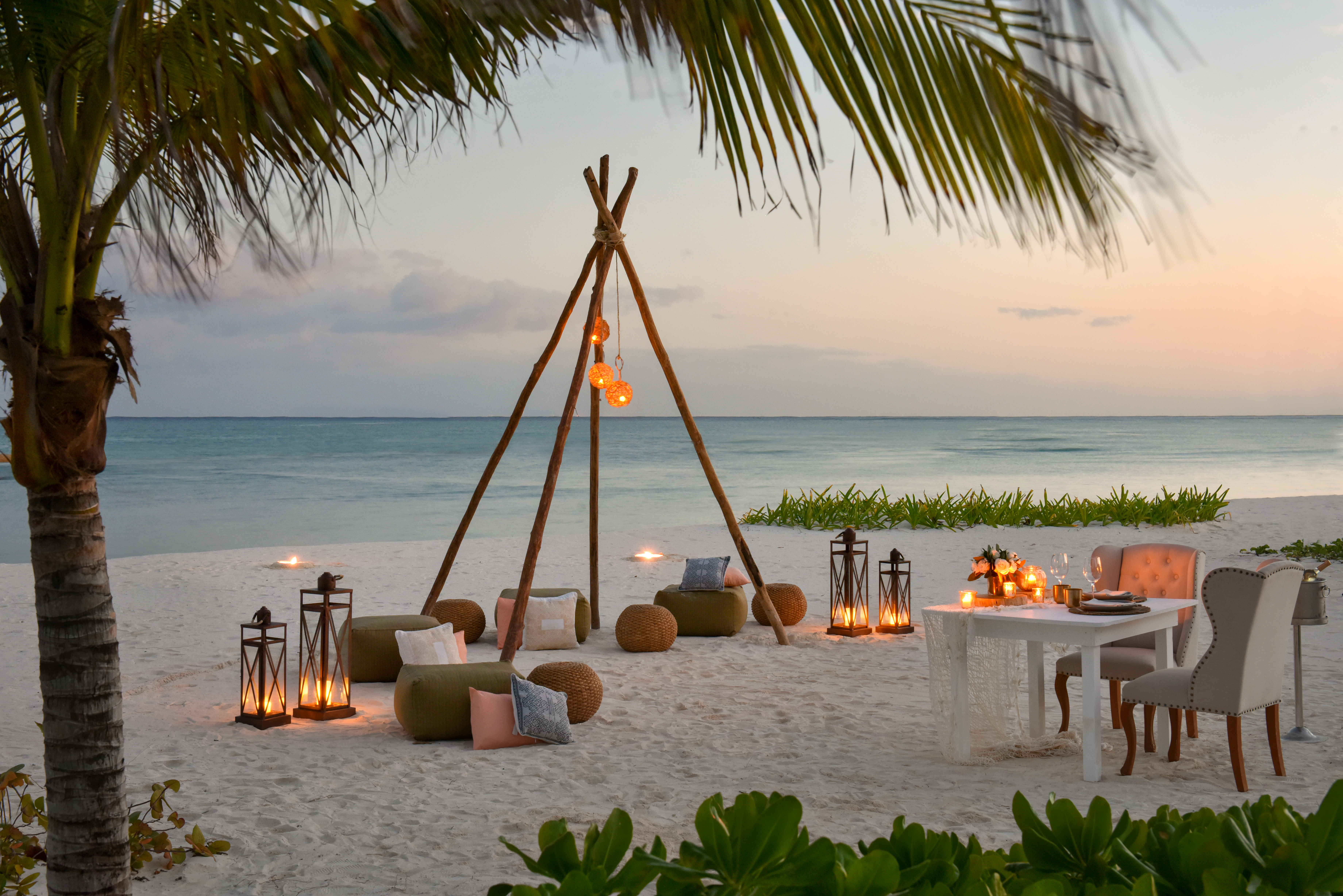 Romantic beach setup with a teepee structure, lanterns, and a table for two on a sandy shore with turquoise water and palm fronds