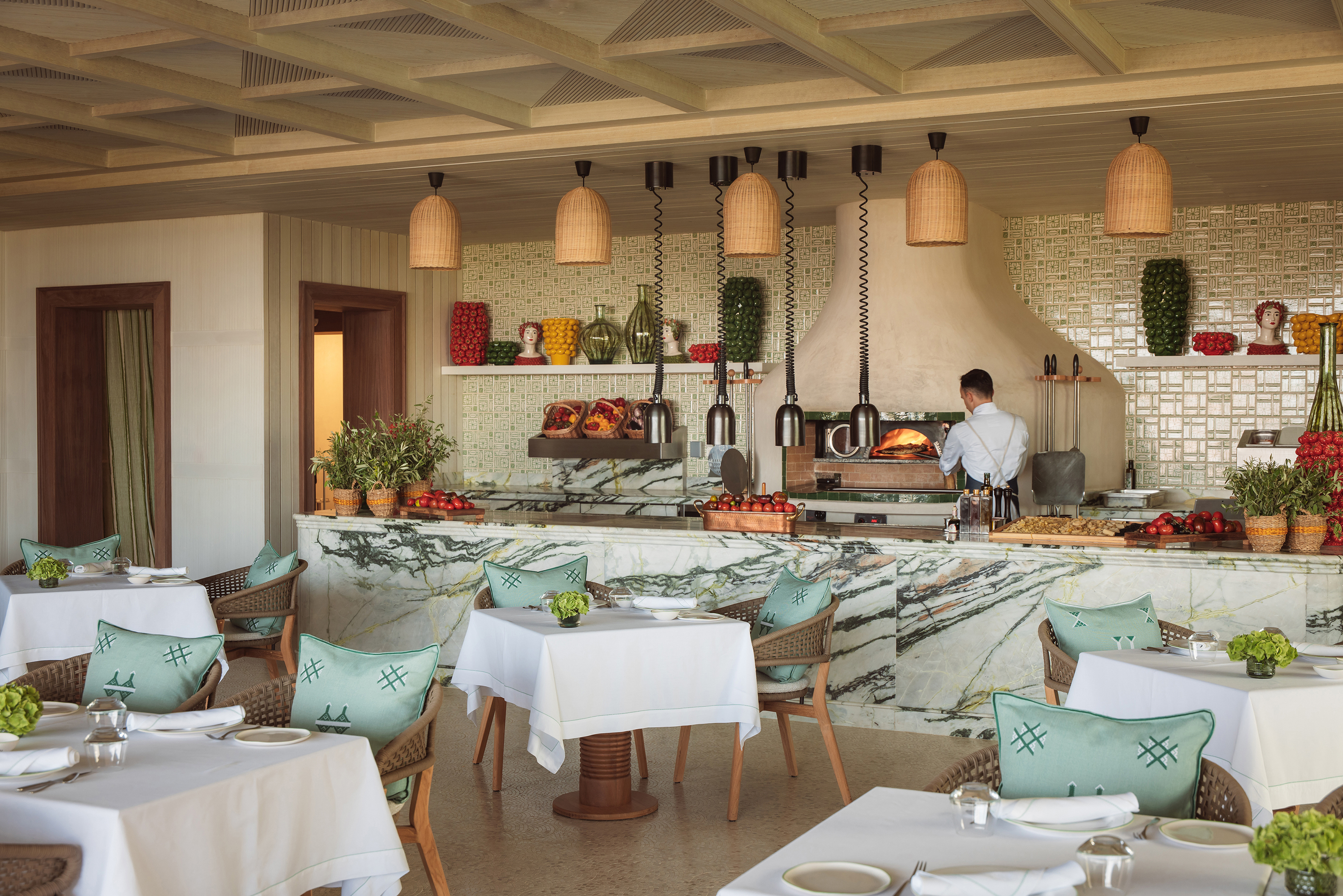 An airy restaurant with tables set to dine and a chef working at a pizza oven in the kitchen area
