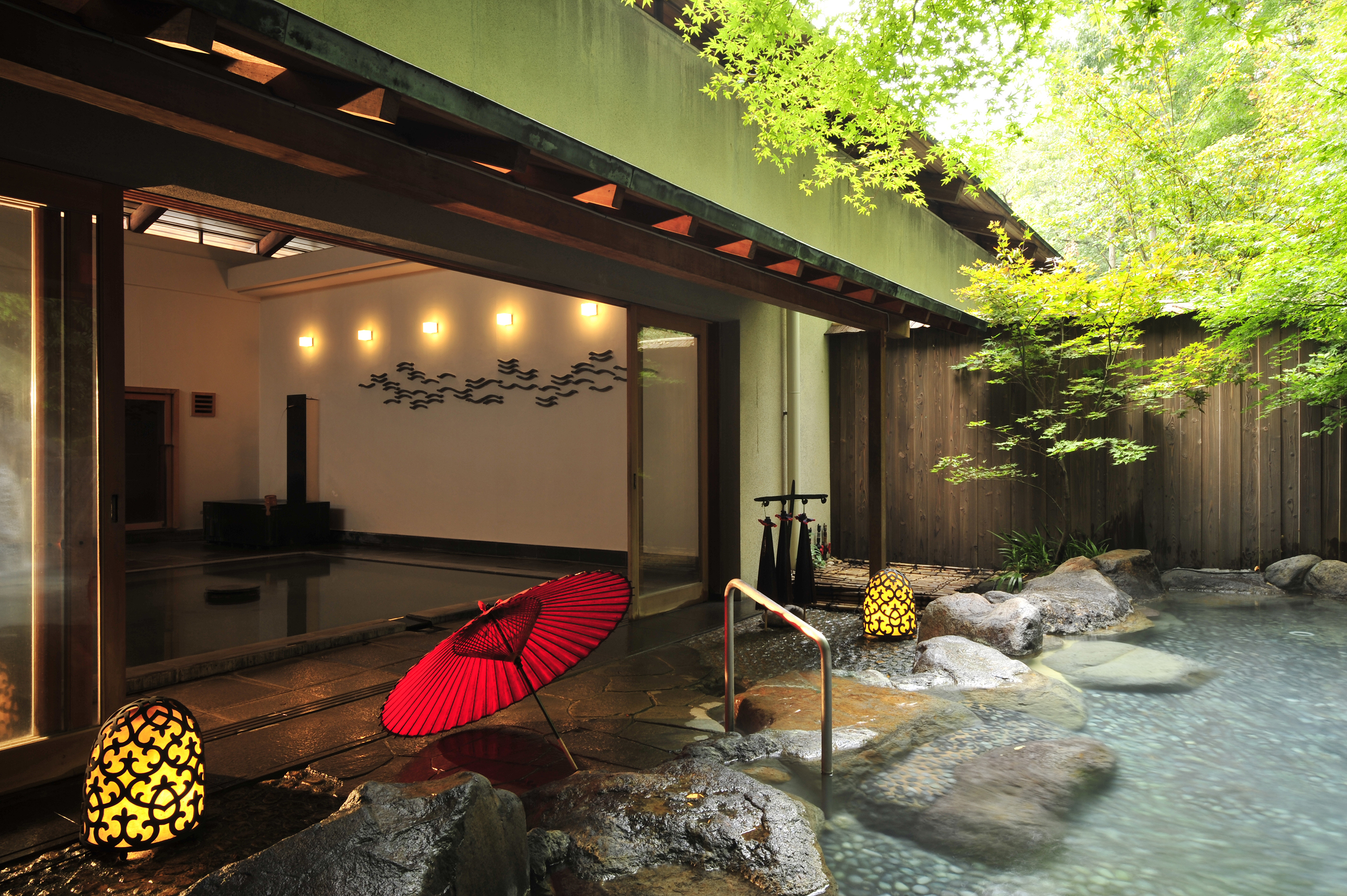 An outdoor onsen with a red umbrella with a glimpse of an indoor pool inside a building beside it