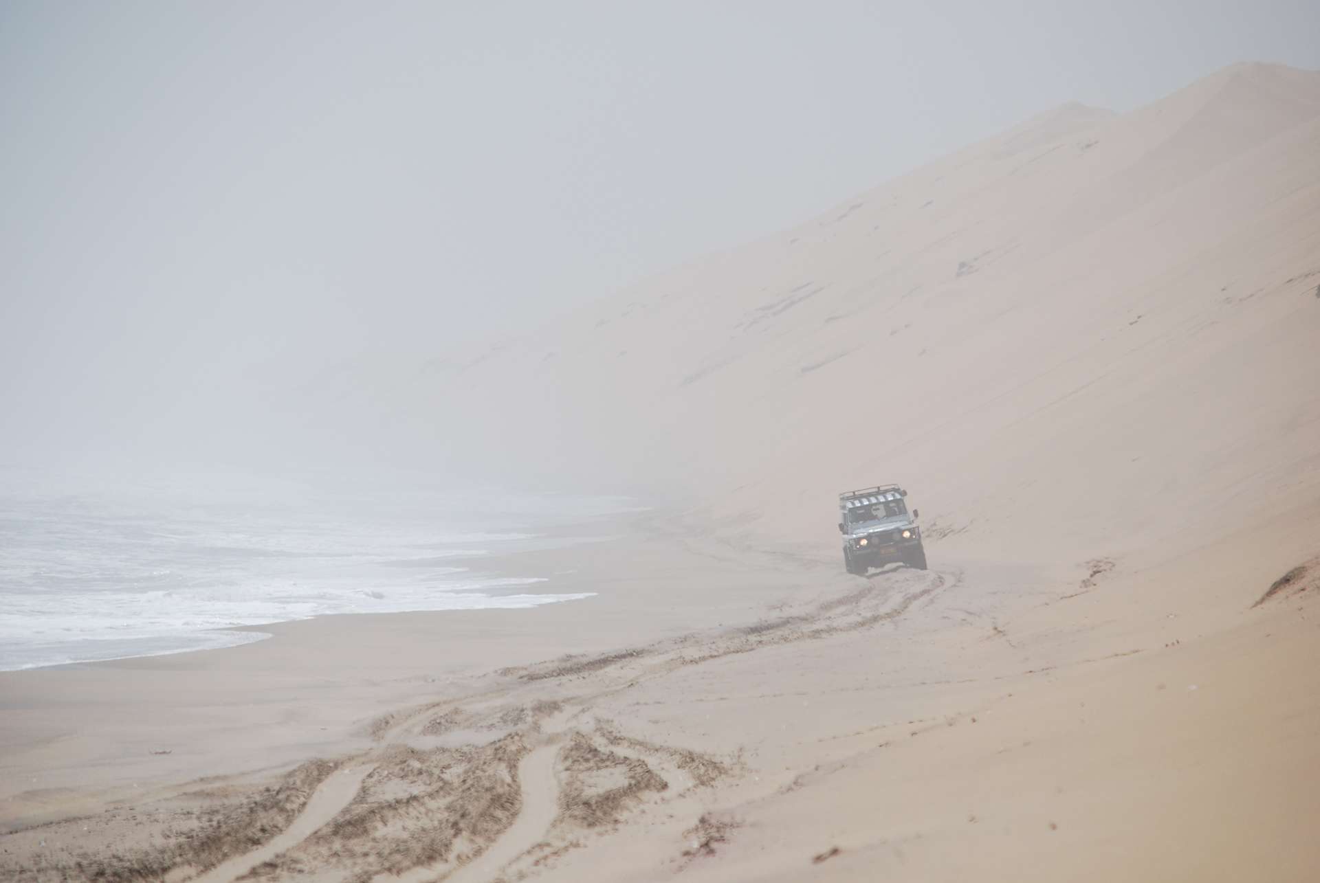 A truck driving on a sandy route in between a tall dune and the sea
