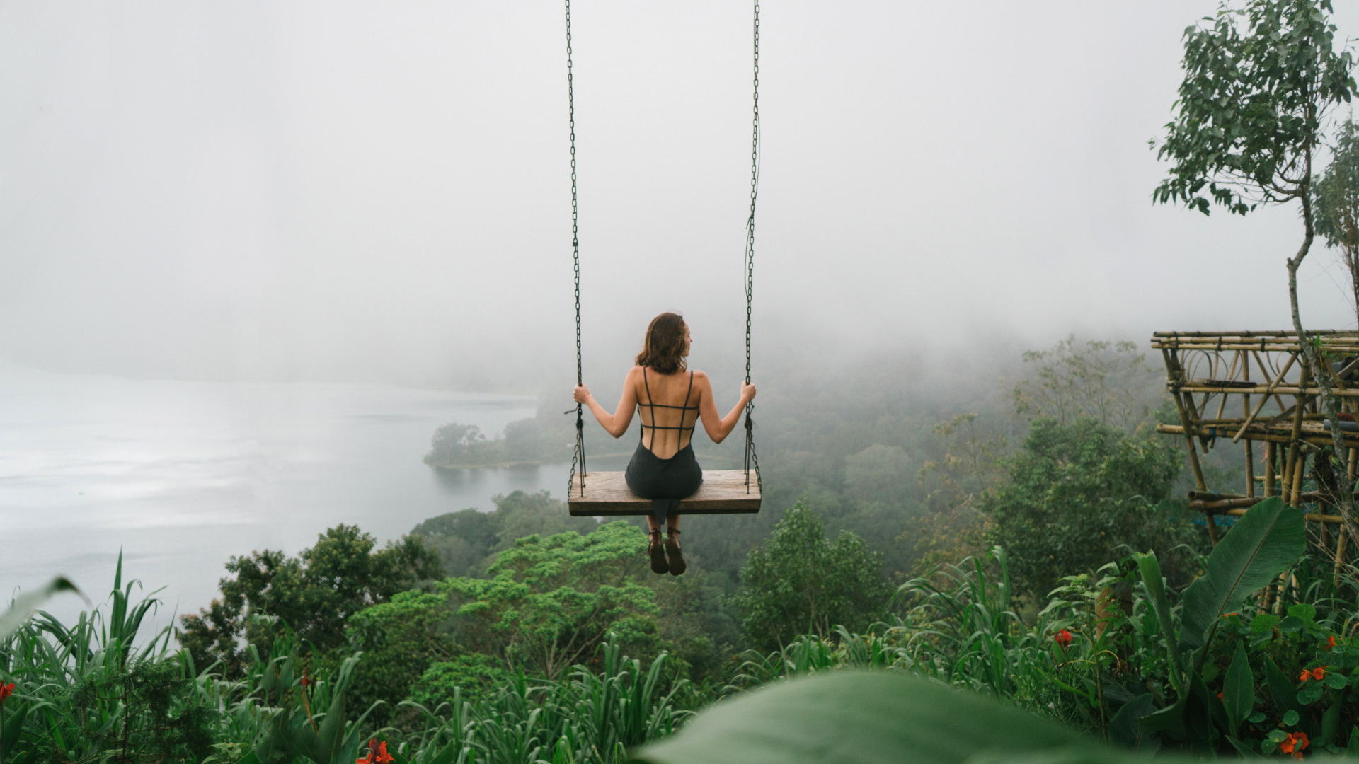 A woman with her back to the camera sits on a swing over a green and misty landscape in Bali
