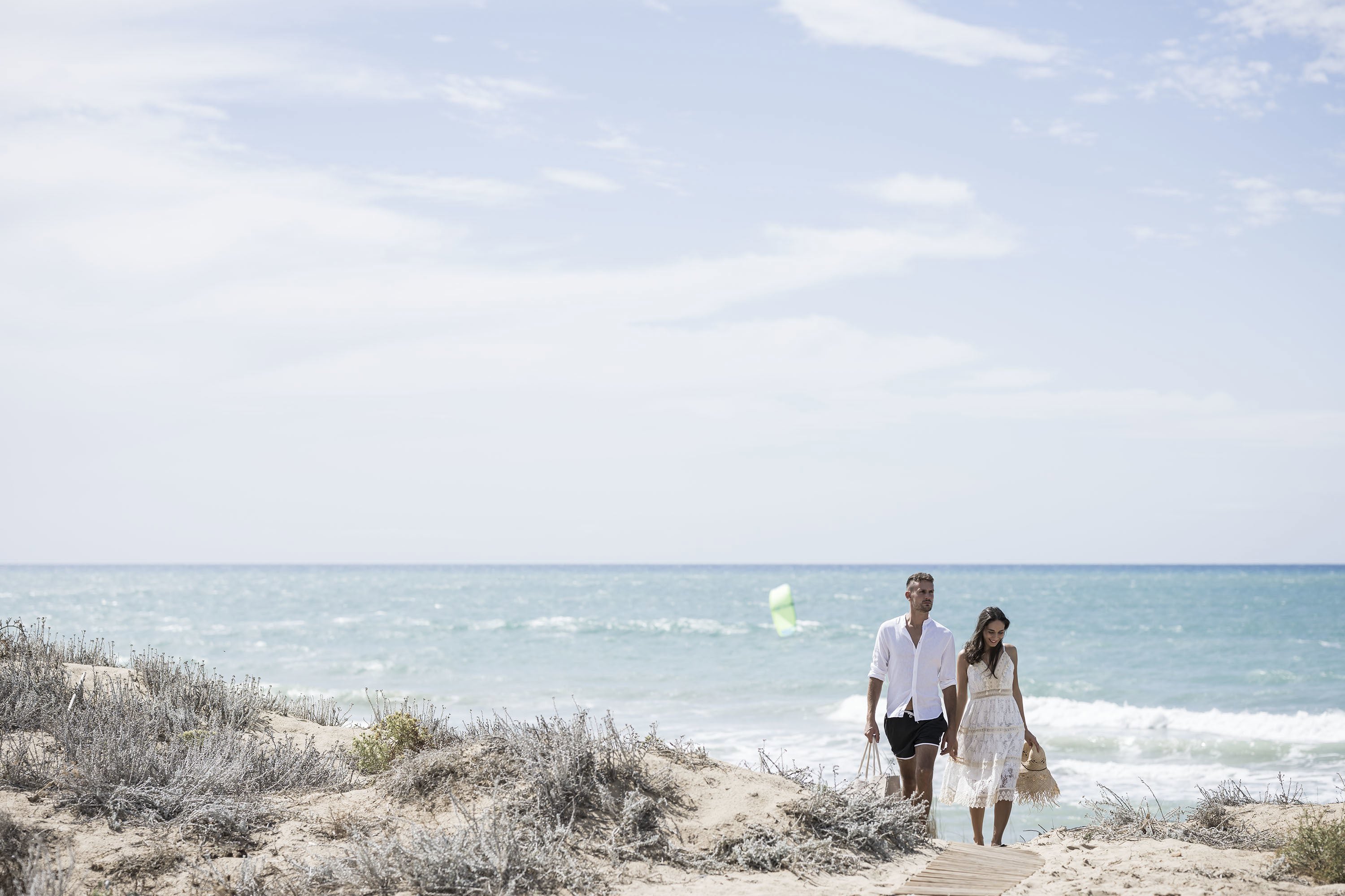 Couple walking hand in hand up a soft sand beach with a kite surfer on the sea in the background