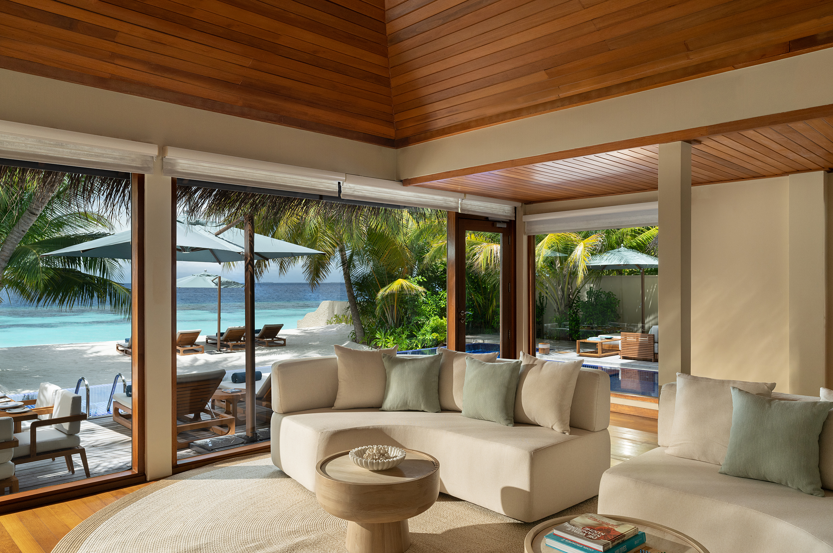 The lounge of a Beach Bungalow at Huvafen Fushi featuring cream sofas and a circular coffee table in front of large windows looking out to a pool and sun loungers