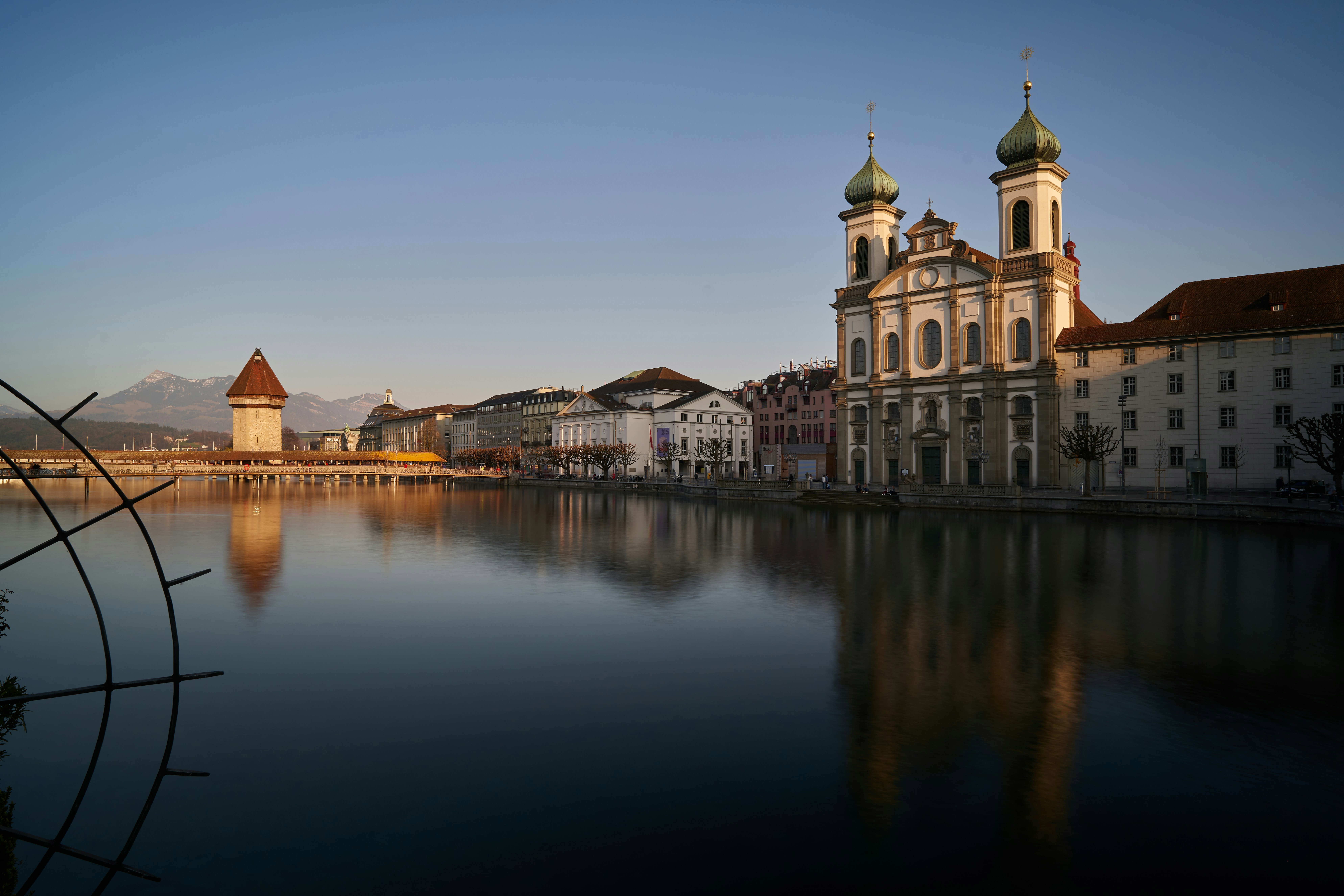 A calming view of the Jesuit Church and Water Tower on the Reuss River in Lucerne, Switzerland, with Mount Pilatus in the background during twilight.
