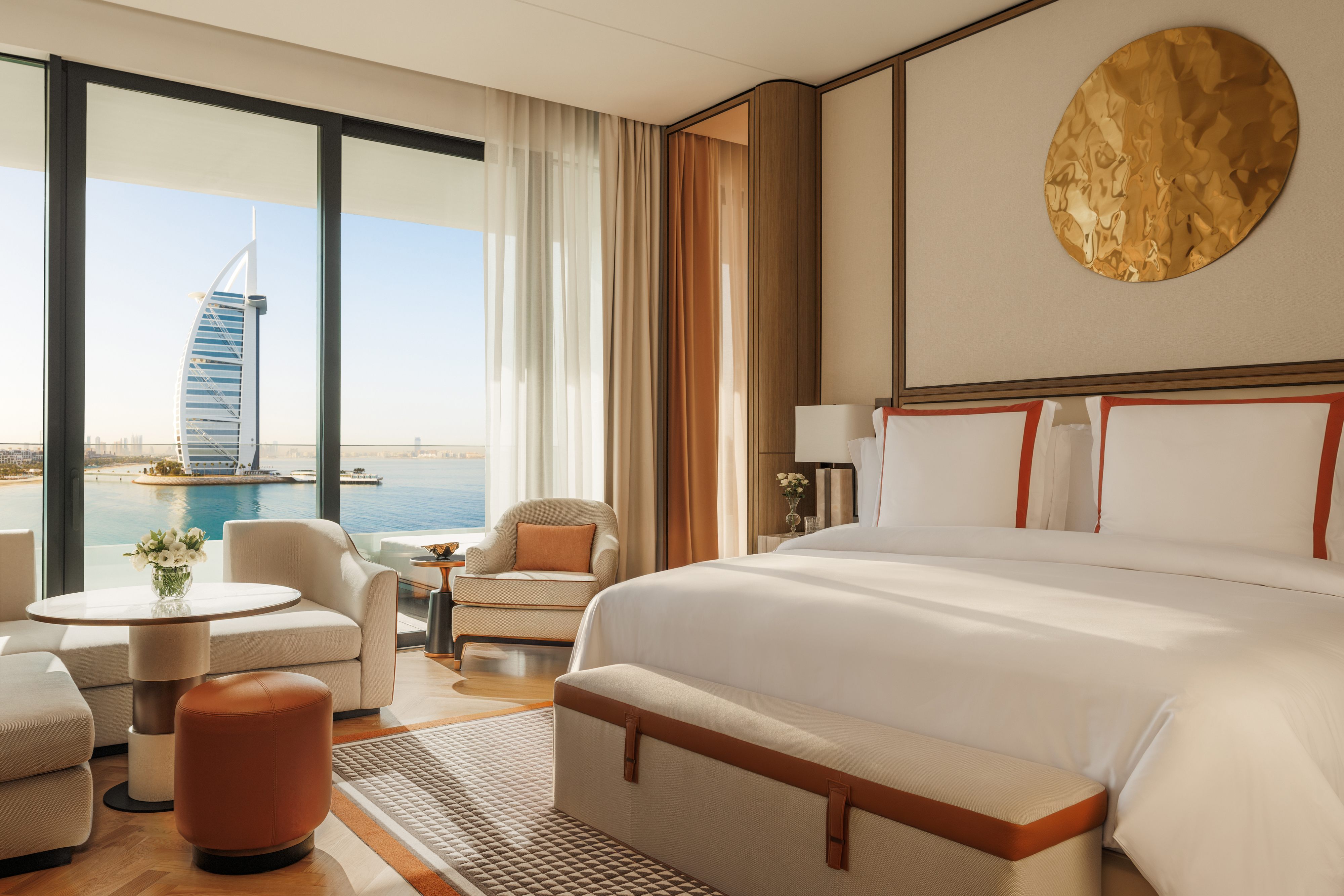 A stylish room decorated in cream and red tones with a bed and comfy seating and a view of the Burj Al Arab