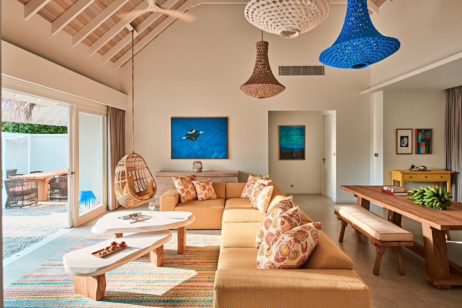 An airy coastal living space in a Two Bedroom Beach Villa at Finolhu featuring a long sofa with patterned cushions, a hanging chair, dining table, coffee tables and decor with blue accents