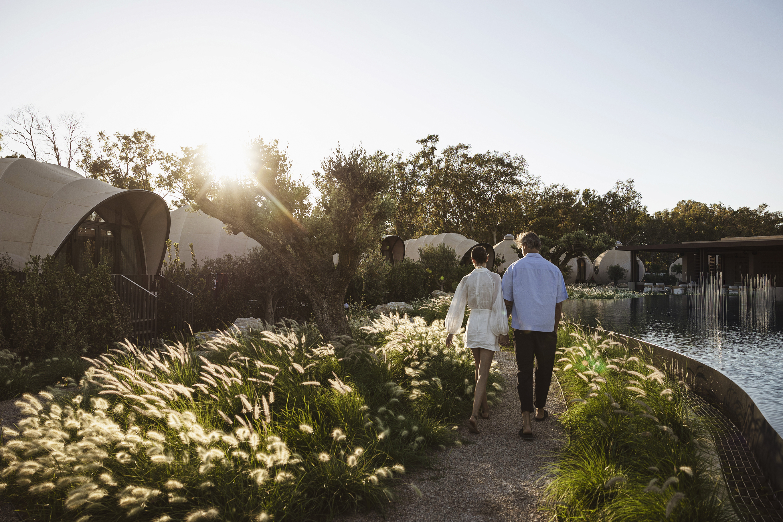 Couples in white and blue strolling through lakeside gardens in front of luxury tents