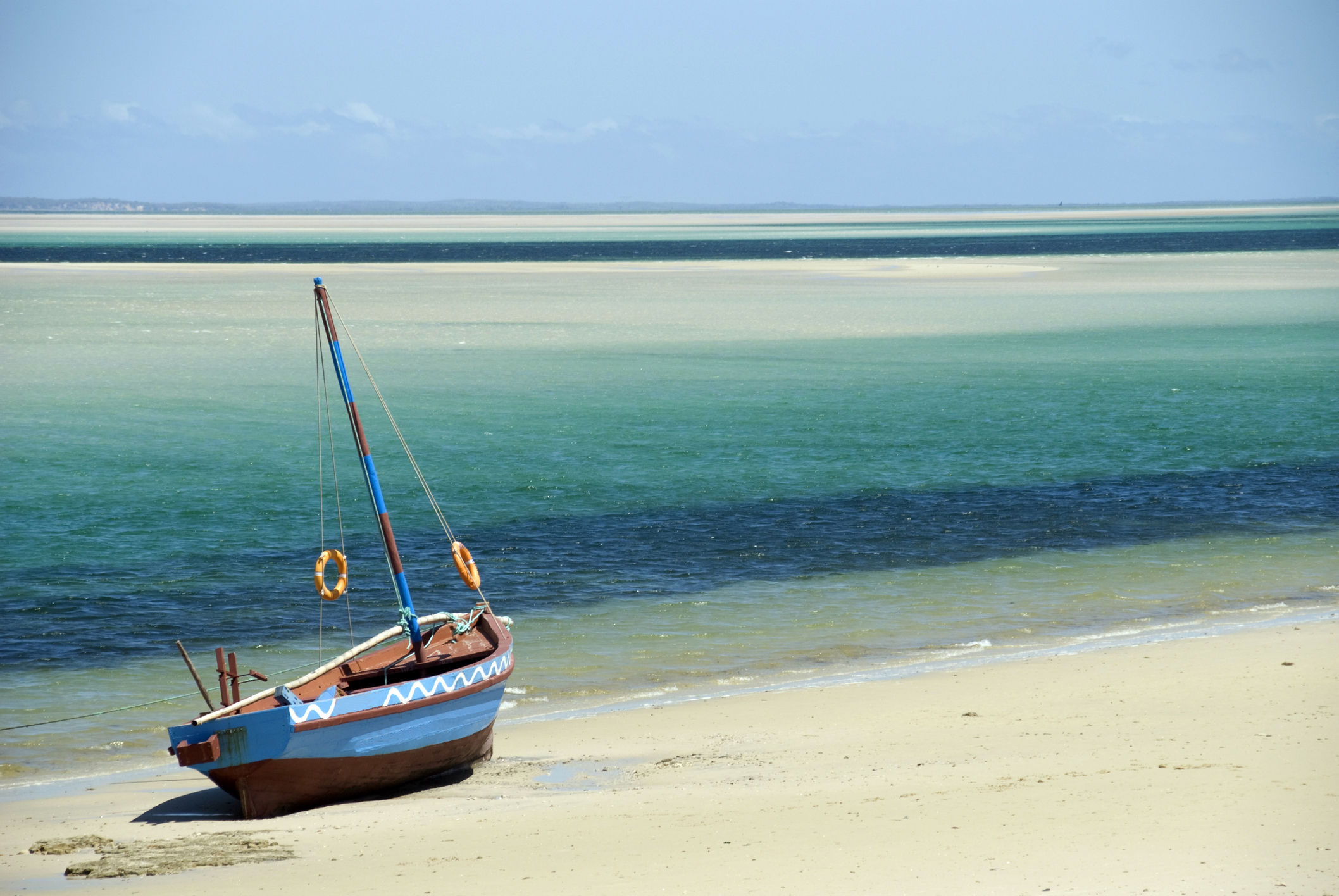 A traditional wooden boat with blue and white detailing is beached on a sandy shore, with clear turquoise waters and a sandbar visible in the background under a blue sky.