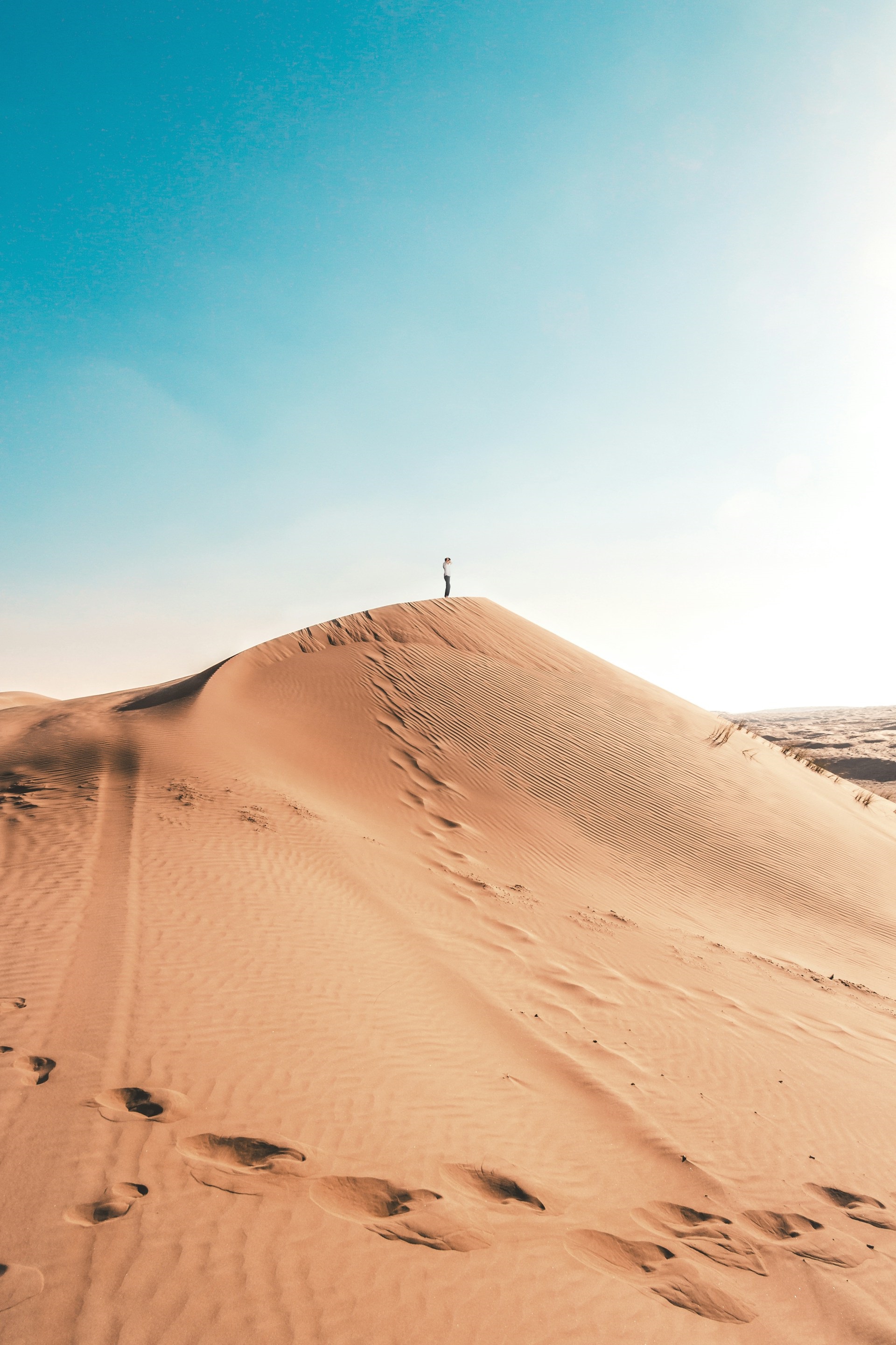 A person stands on top of a sand dune in a desert with footprints leading up to the peak under a clear blue sky.