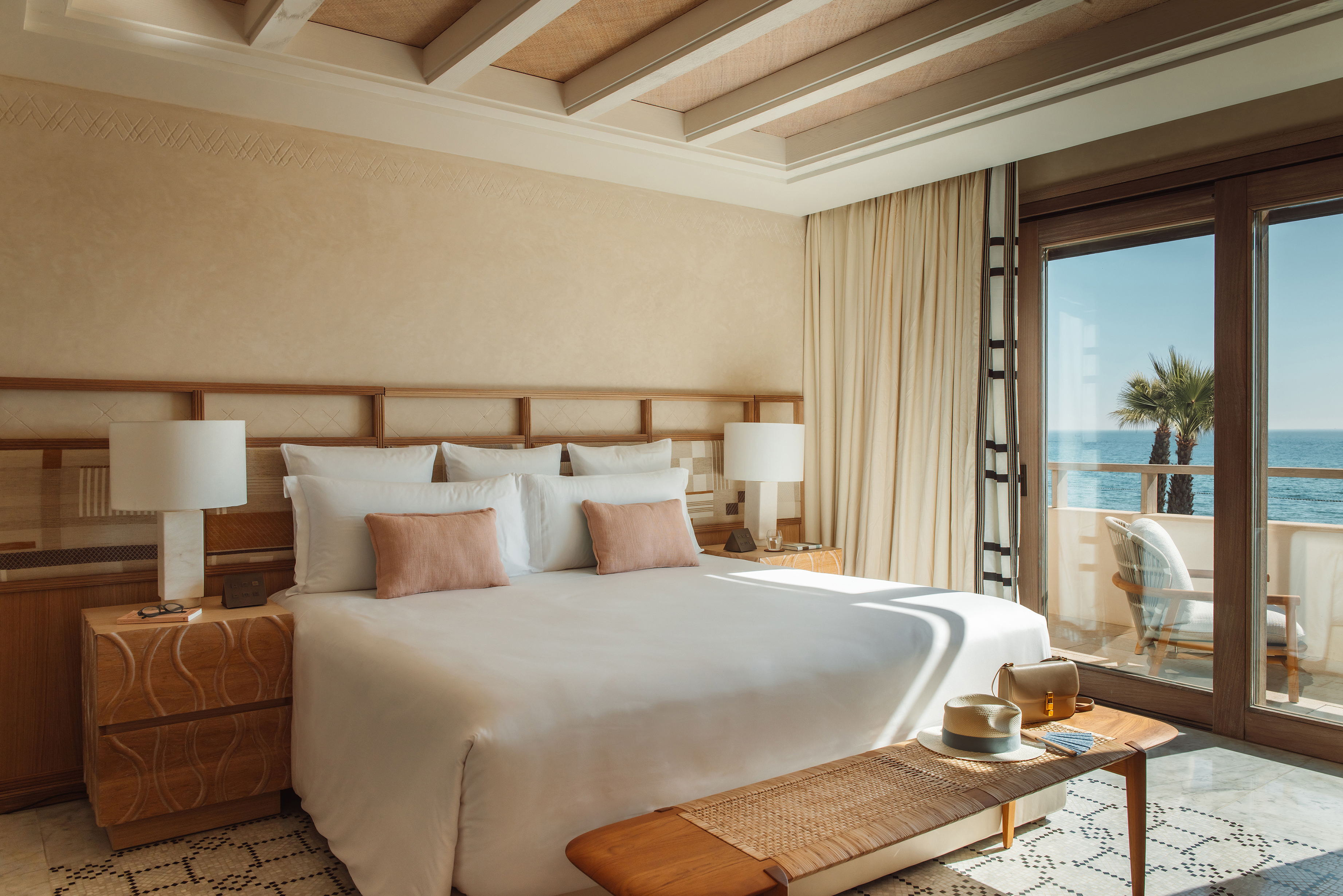 A comfy suite in neutral tones featuring a bed flanked by lamps and wooden headboard, with a sunny terrace outside