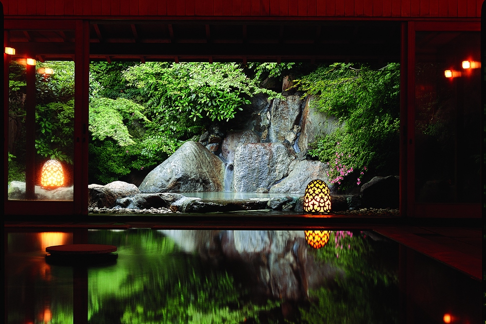 An indoor onsen bath with a view of an outdoor onsen surrounded by rocks through sliding doors