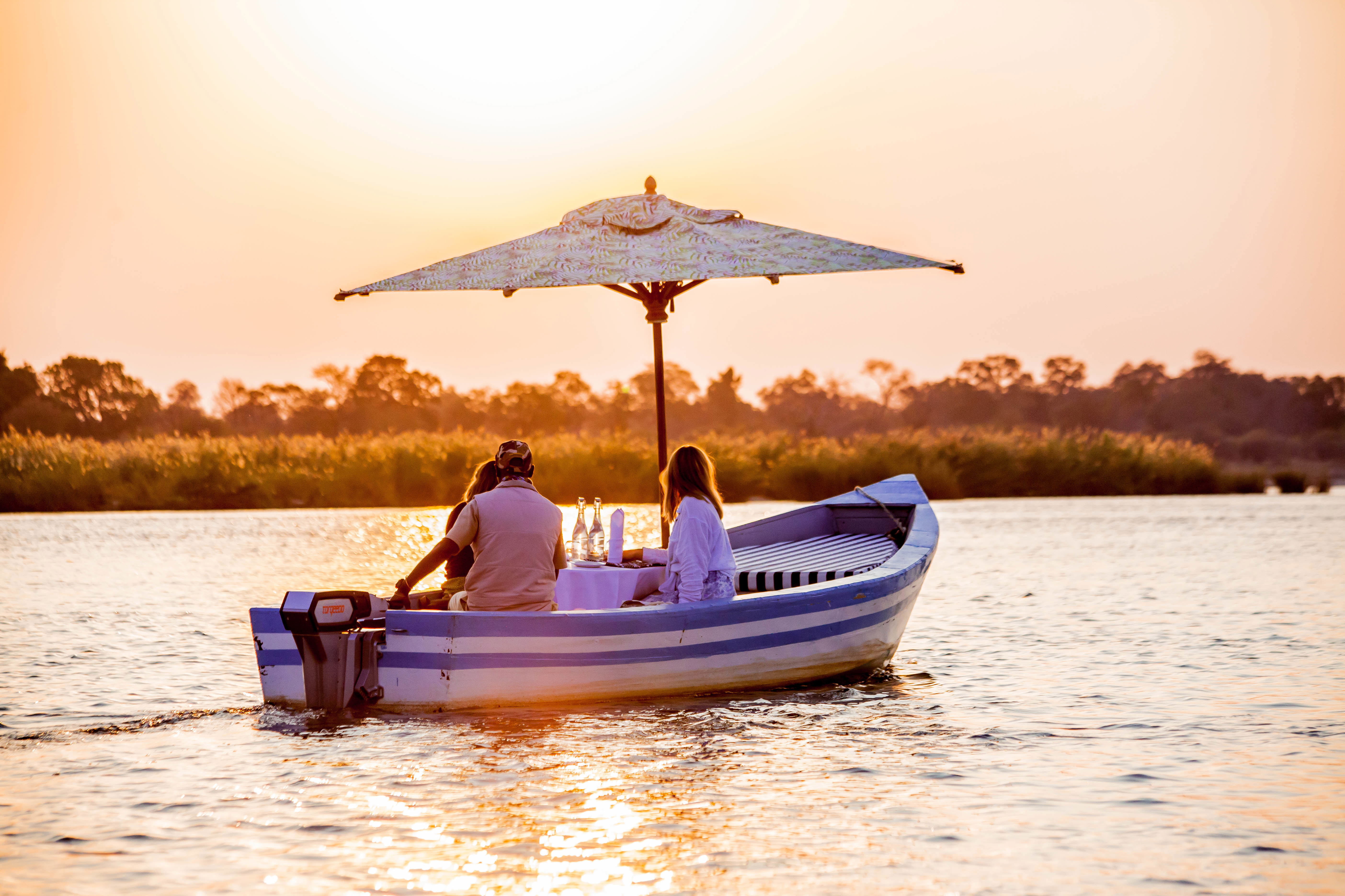 Couple and a guide enjoying a sunset boat ride on the Zambezi River with an umbrella for shade.