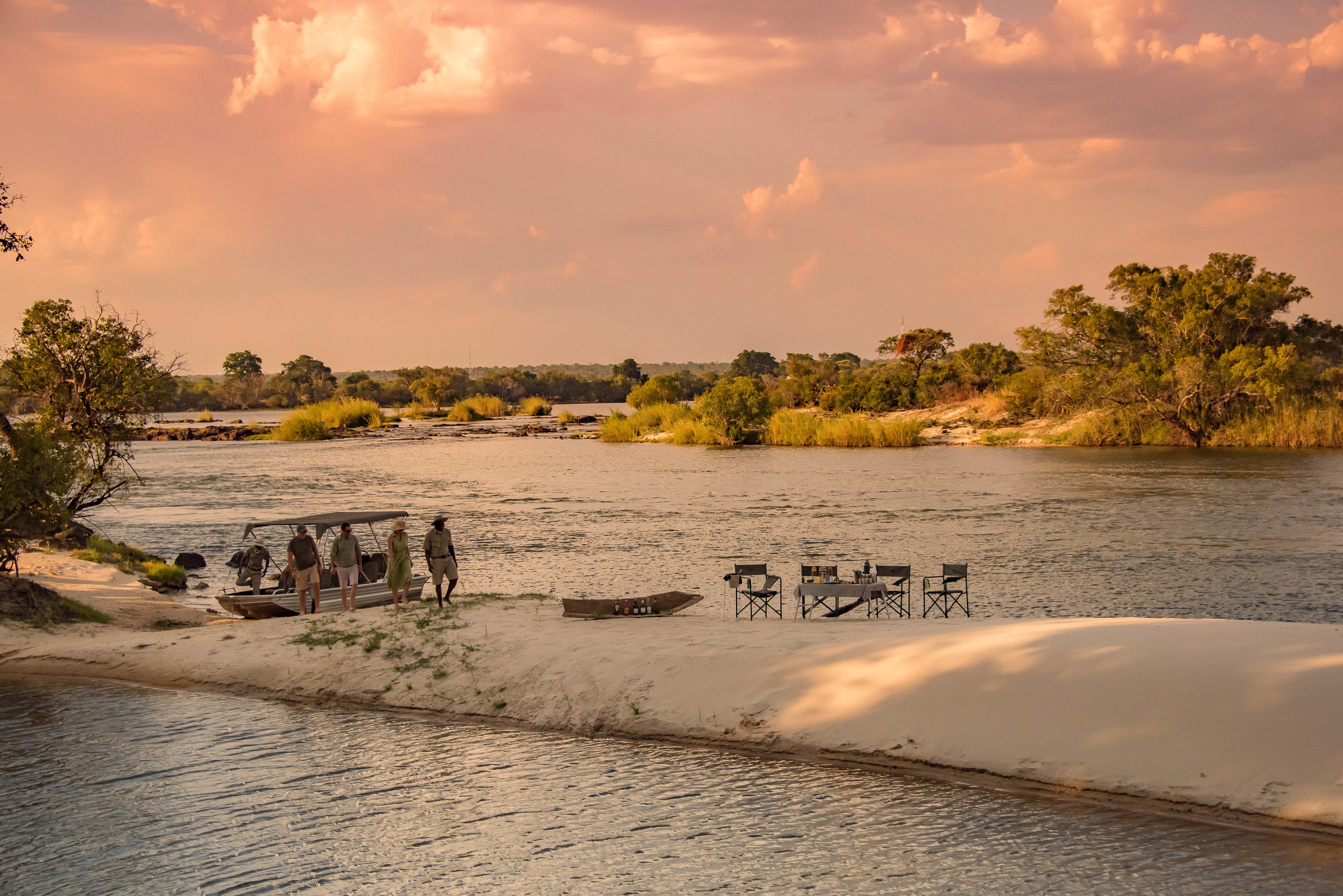 A group of people walking from a boat to a seating area on a sand bank along the Zambezi river