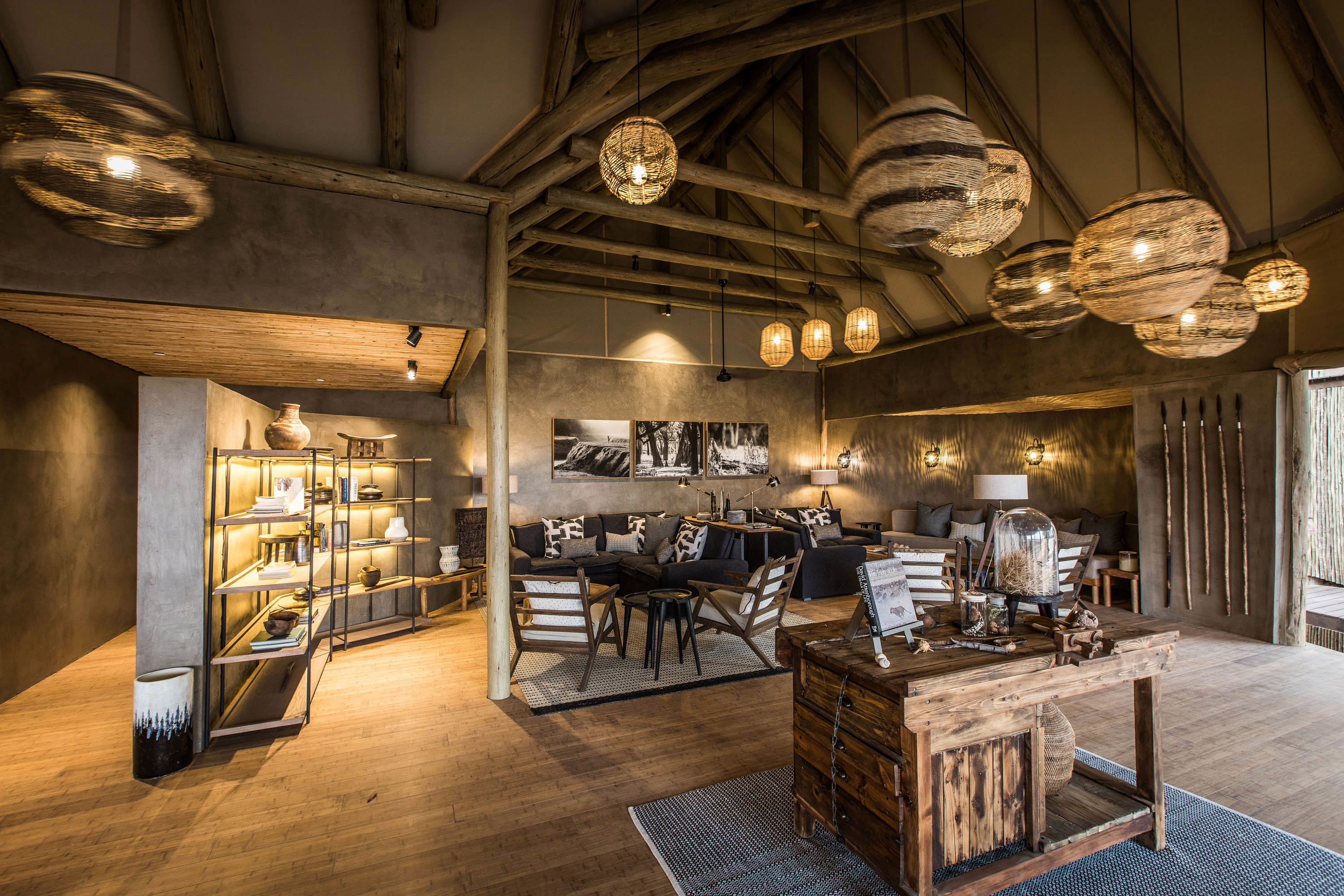 Lounge of Puku Ridge Camp featuring a rustic, open-plan area with wooden beams, wicker light fixtures, and comfortable seating, including a central wooden table with decorative items and bookshelves on the side