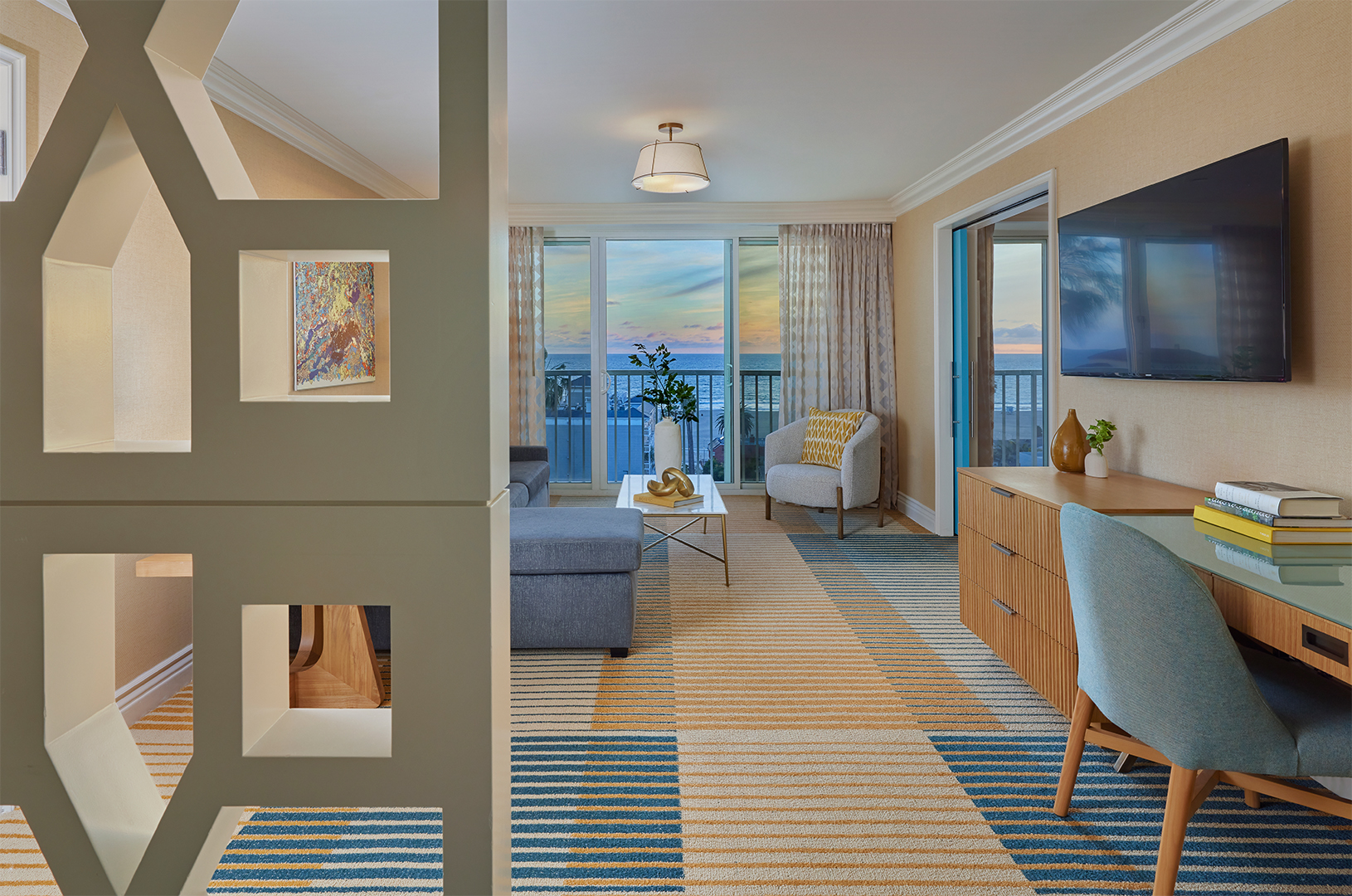 A living room of a hotel suite with a window view of a blue sky. The room is decorated in cream and blue tones and features a desk, sofa and TV area. The view into the room is obstructed by a wall divider.