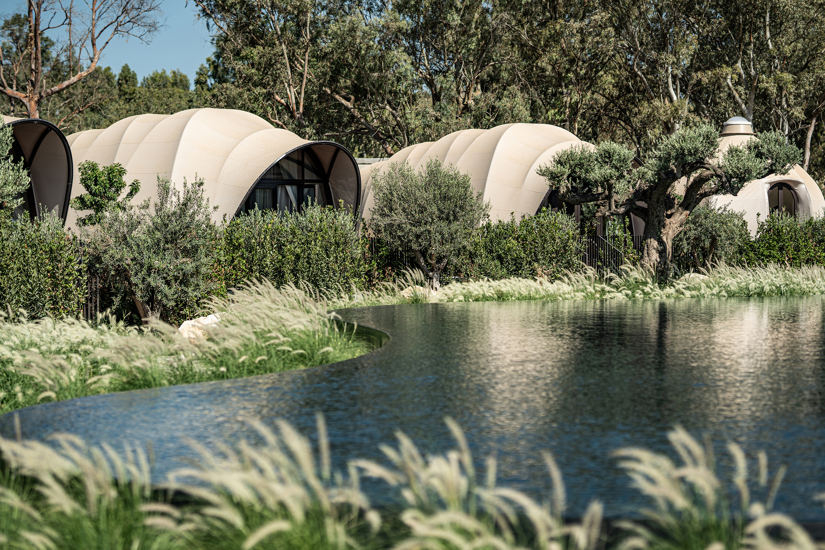 Lake flanked by established greenery hiding domed tented cabanas with glass fronts