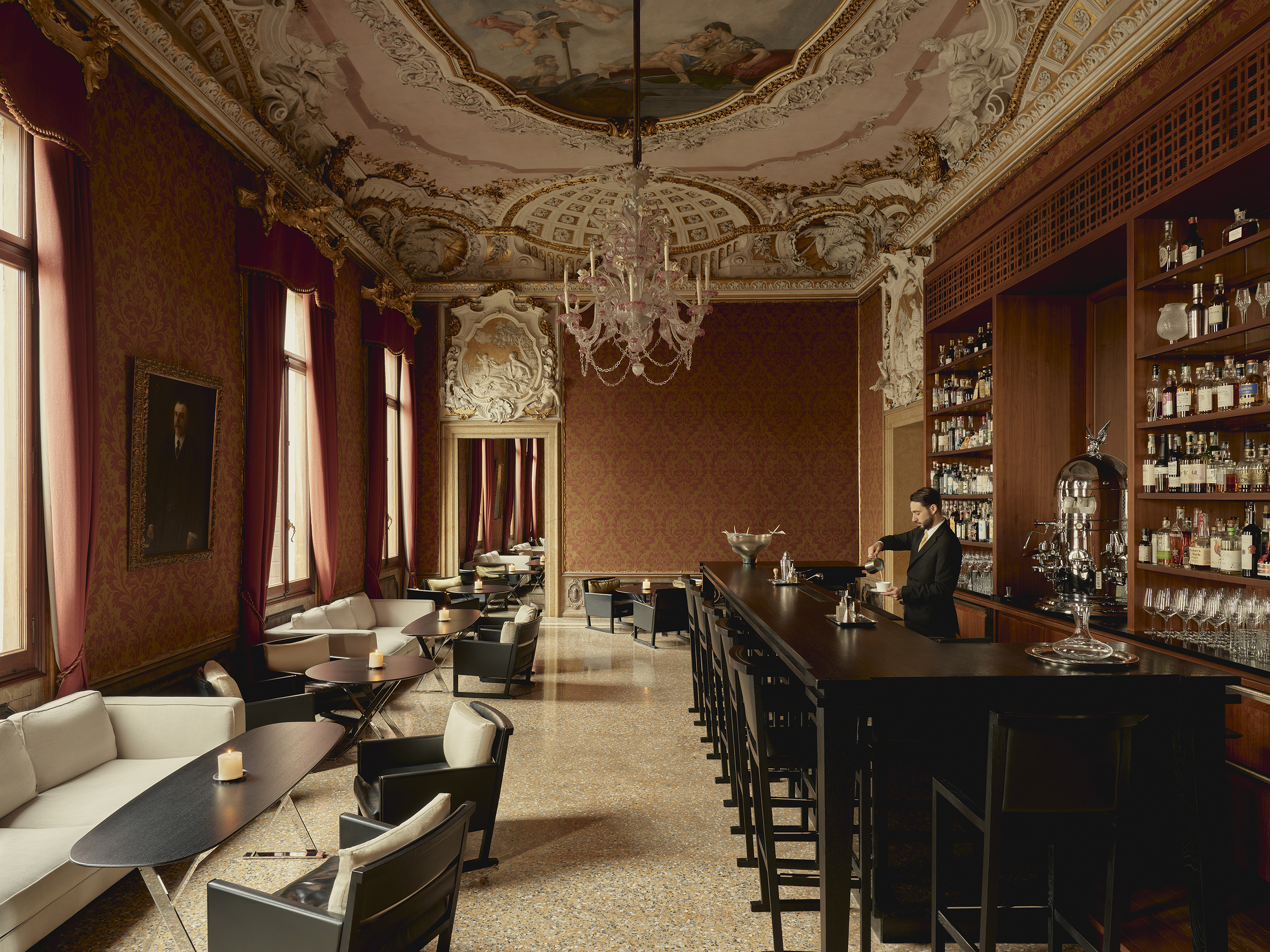 A large bar with detailed fresco ceiling and chandelier above contemporary furniture and a bartender mixing drinks
