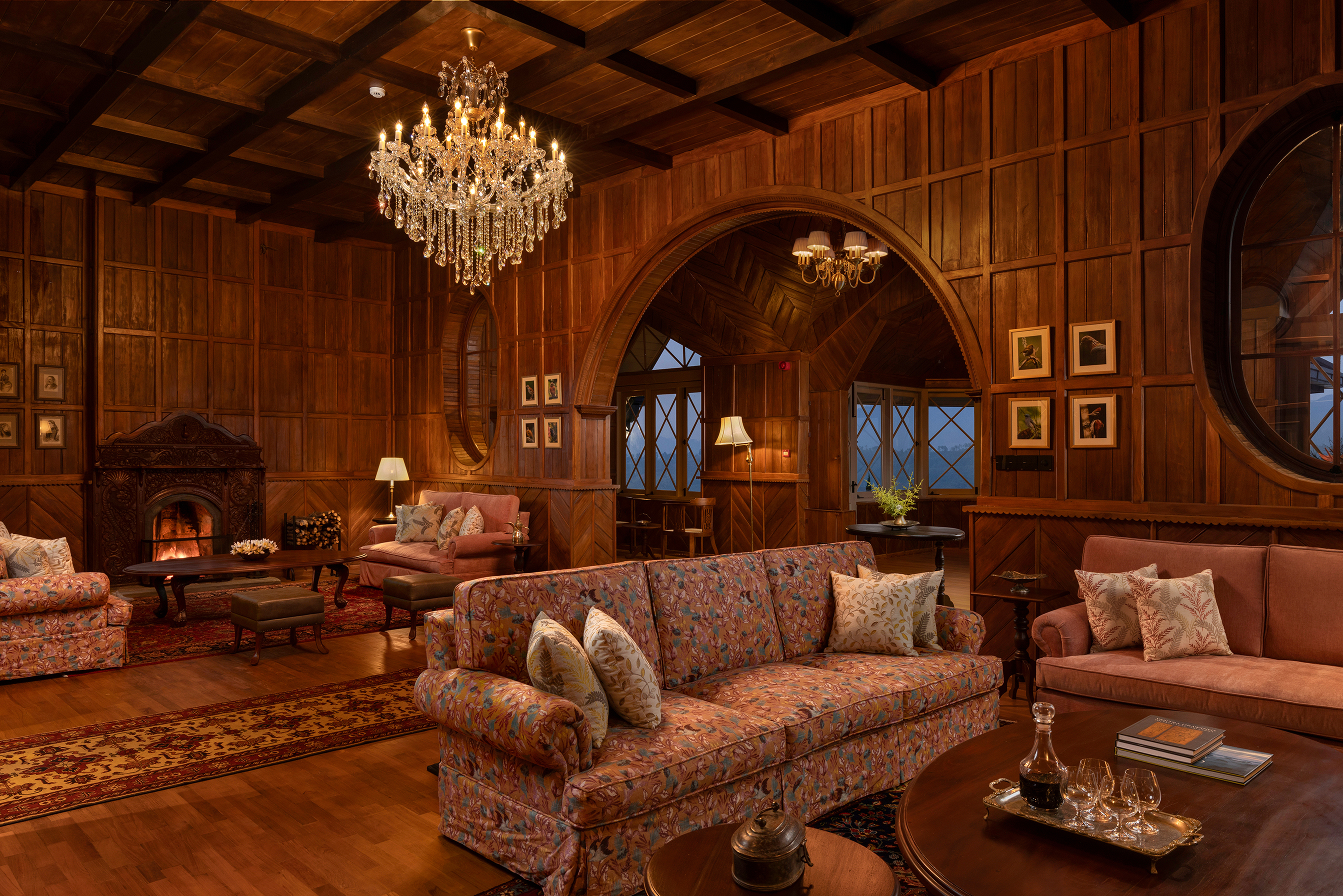 The cosy lounge of Uga Halloowella at night featuring comfy floral sofas, a lit fireplace and wooden panelled walls and archways beneath a chandelier