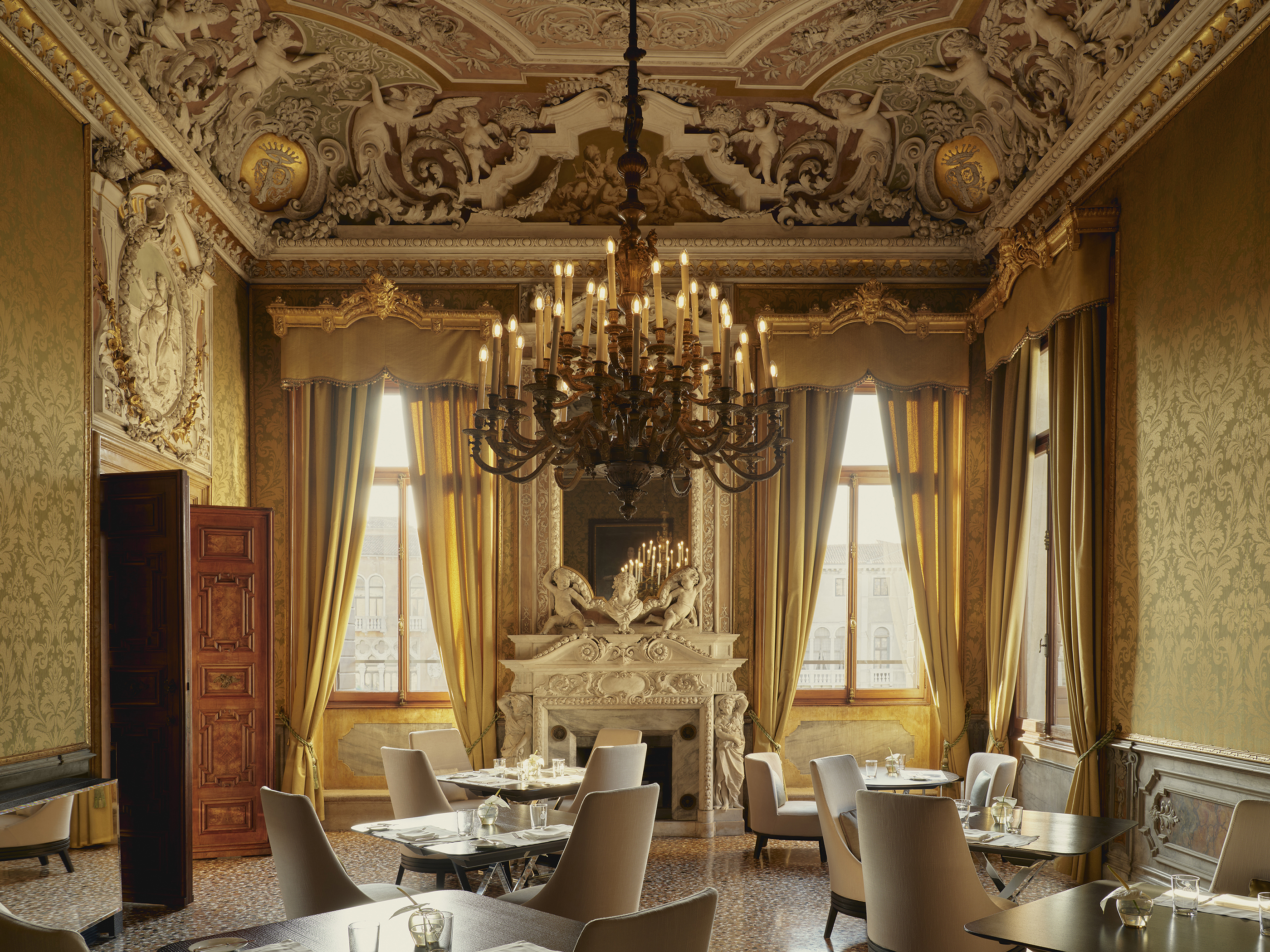 A lavish dining room with ornate carved ceiling and walls featuring a chandelier and large curtains, with sleek white tables set for a meal