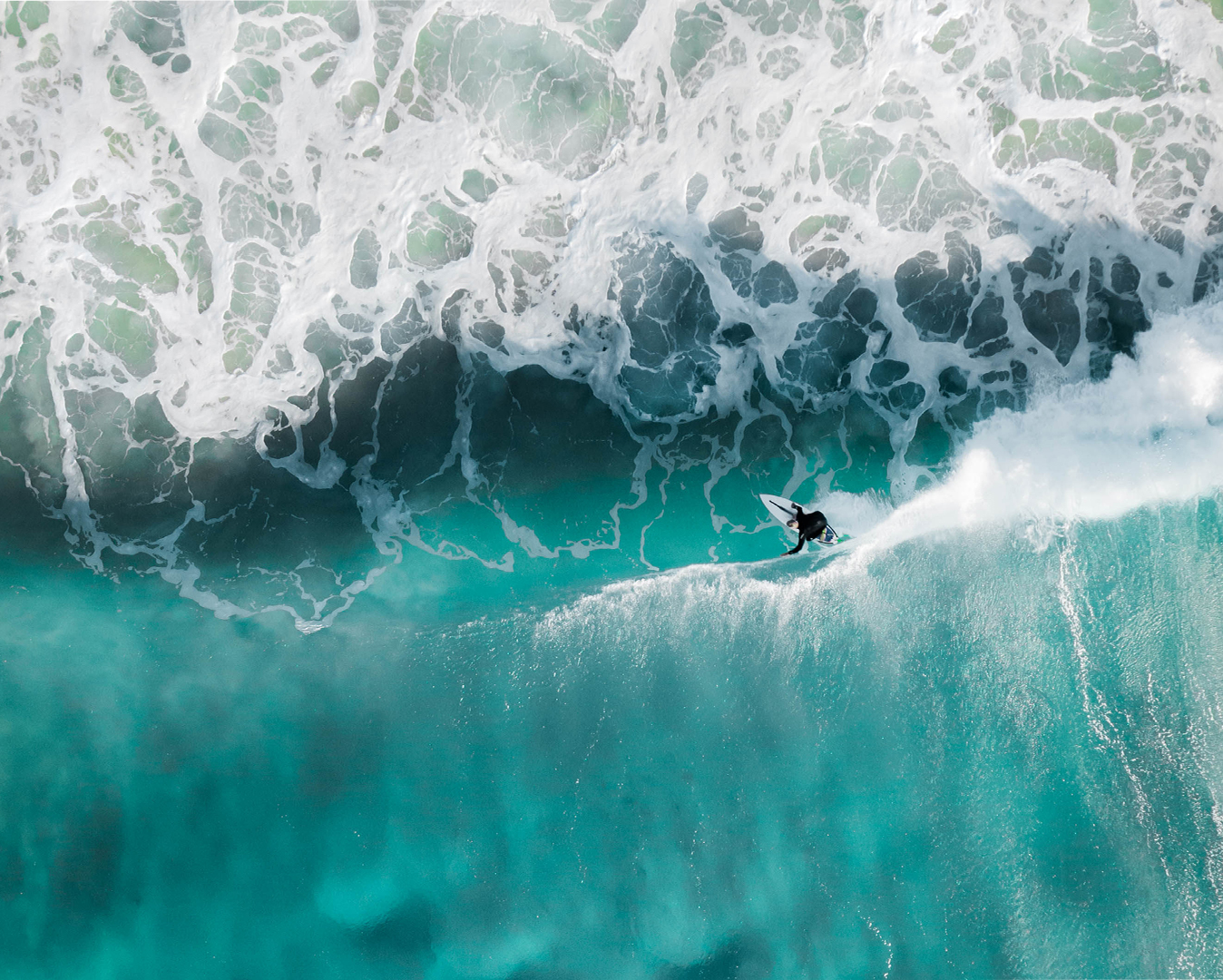 A surfer paddles towards a turquoise wave with white foam from an aerial perspective.