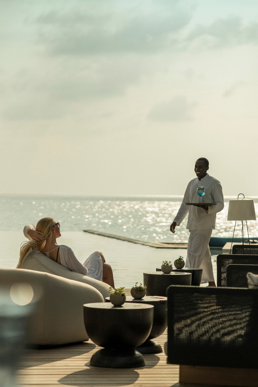 A woman lounging on a pool deck as a waiter brings her a drink