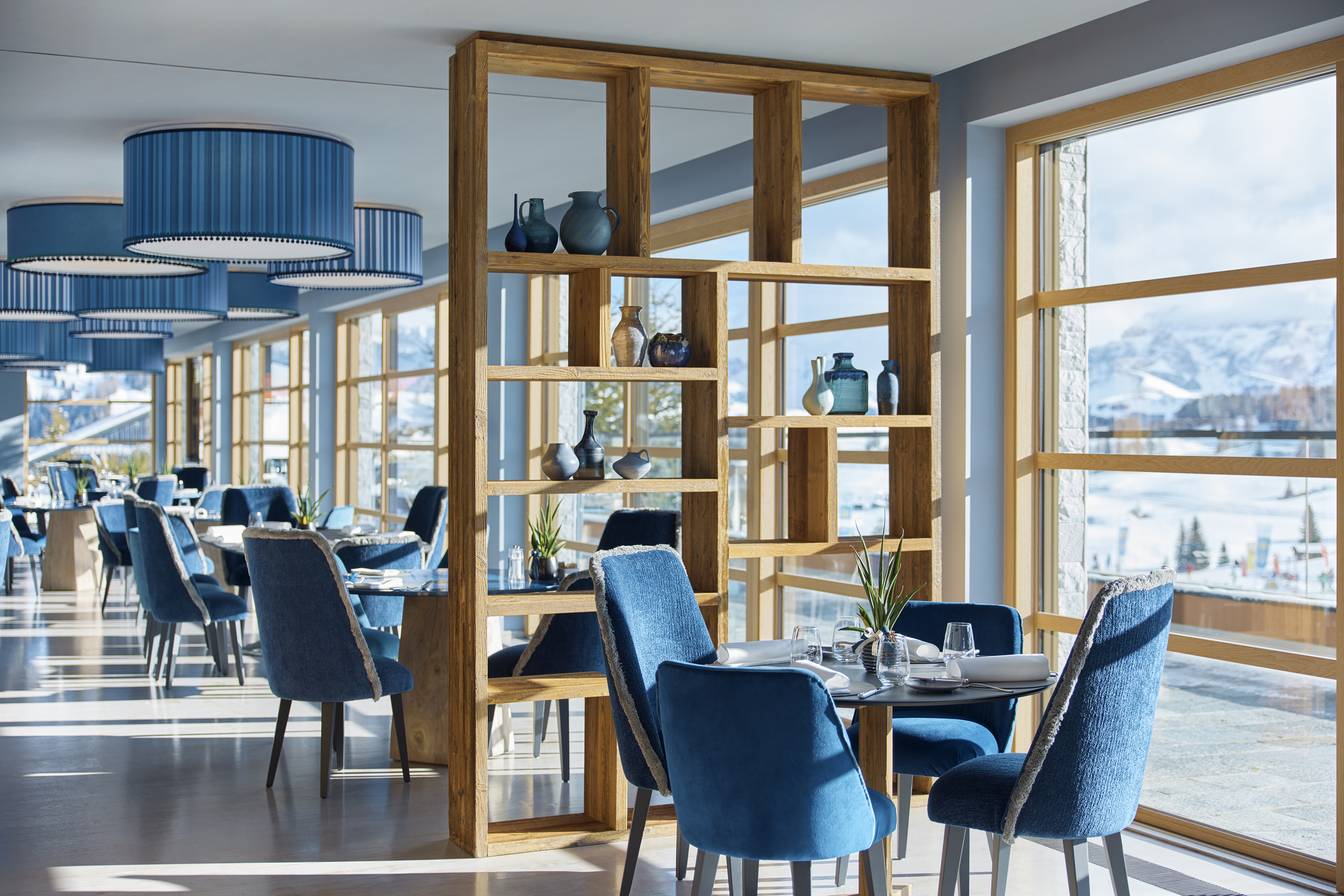 Sassolungo restaurant with modern interior featuring blue upholstered chairs, wooden tables, and shelving units displaying decorative items, with large windows offering panoramic views.