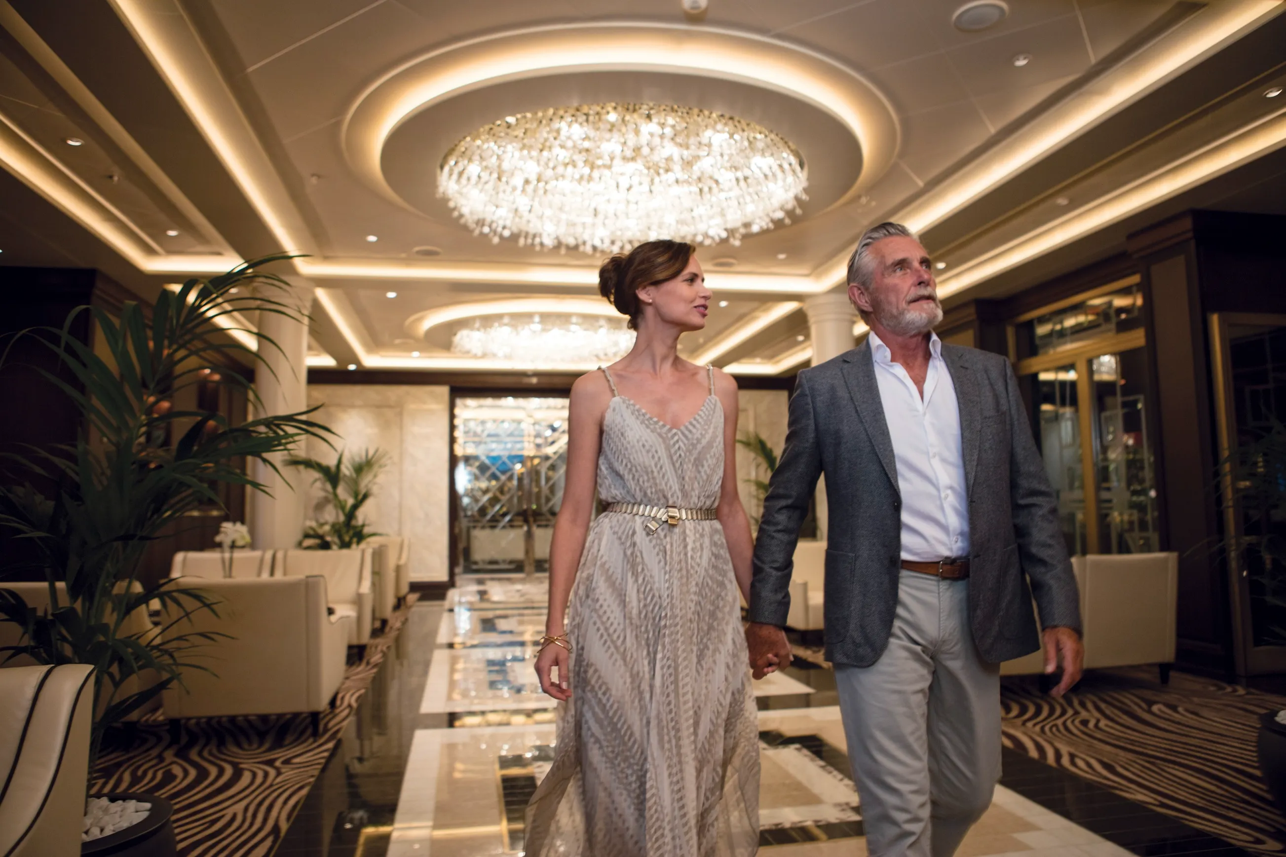 Couple holding hands walking through a chandelier-lit hallway on a cruise ship with elegant decor and seating areas.