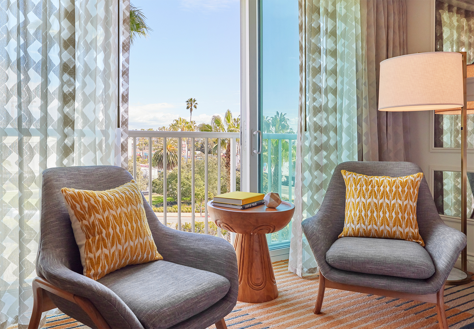 A balcony nook in a hotel suite. The French windows open to a view of palm trees and blue skies. Two blue armchairs and a small wooden table sit in front of the windows.