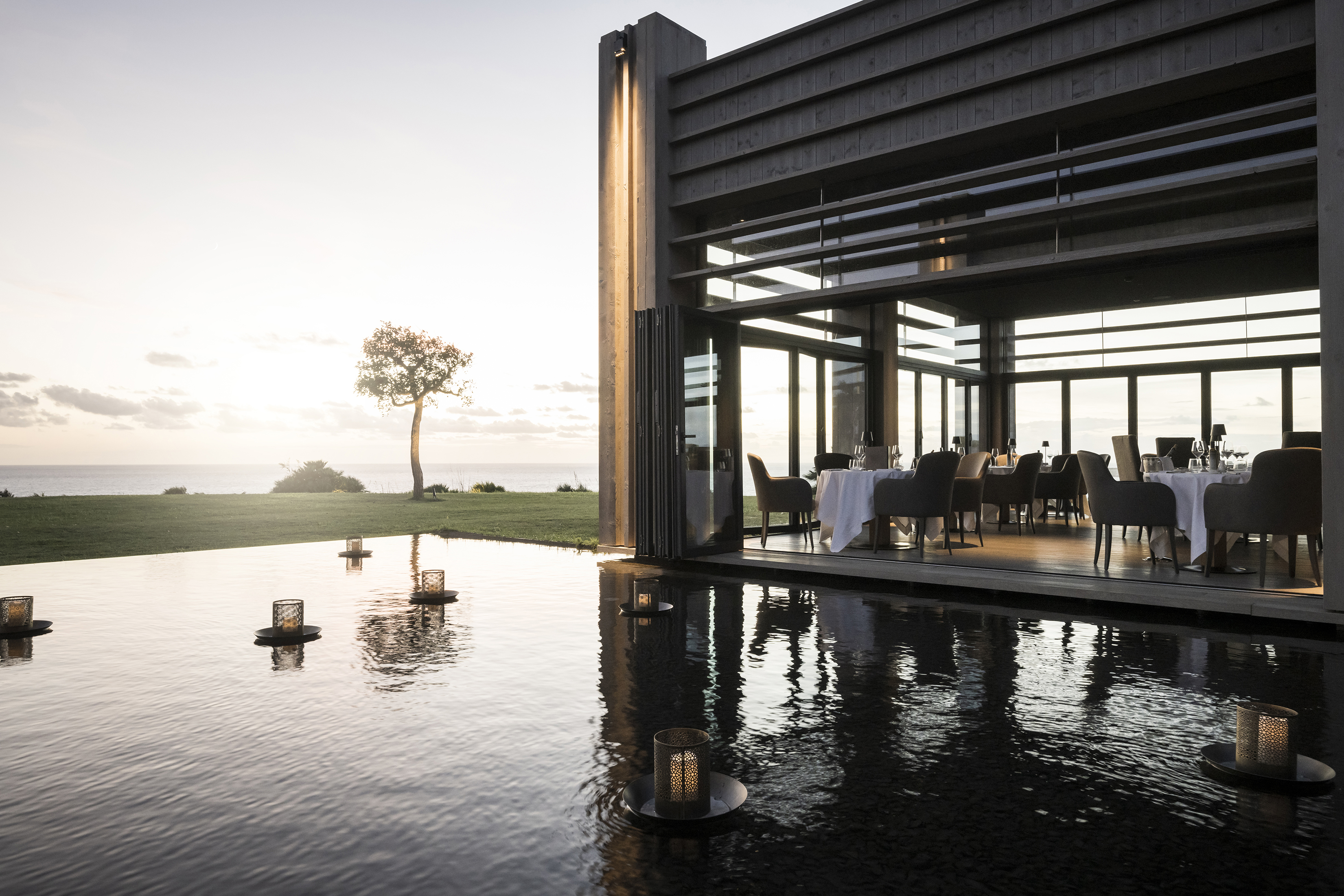 Glass-fronted restaurant set up for dinner beside a pool with floating lanterns and a singluar tree behind 
