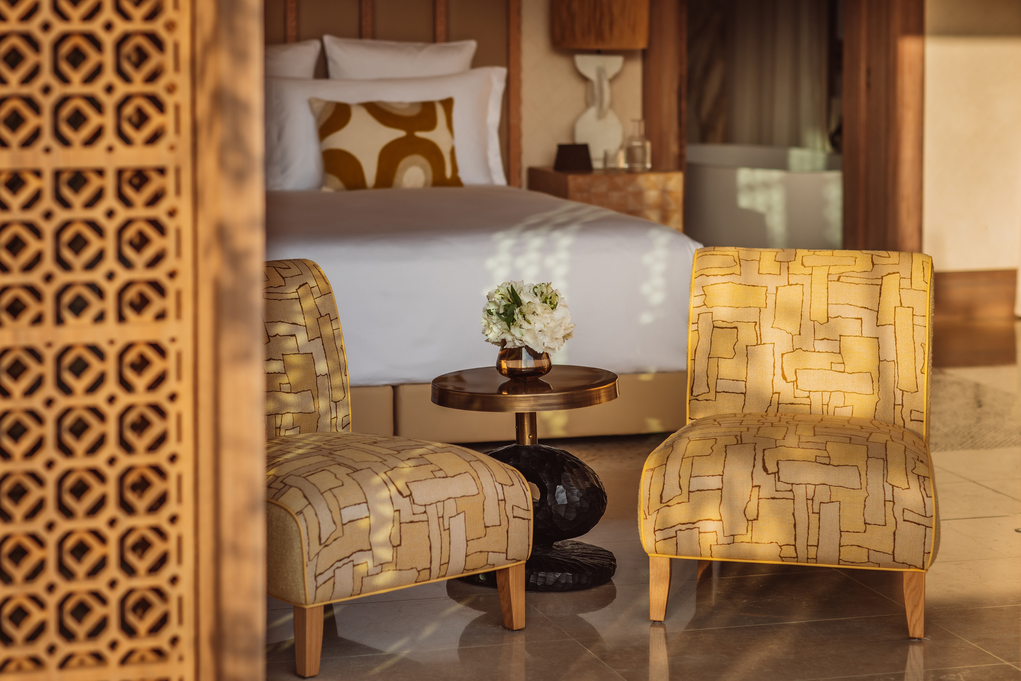 Two patterned chair flanking a small table with a flower and vase, with a glimpse of a bed in the background