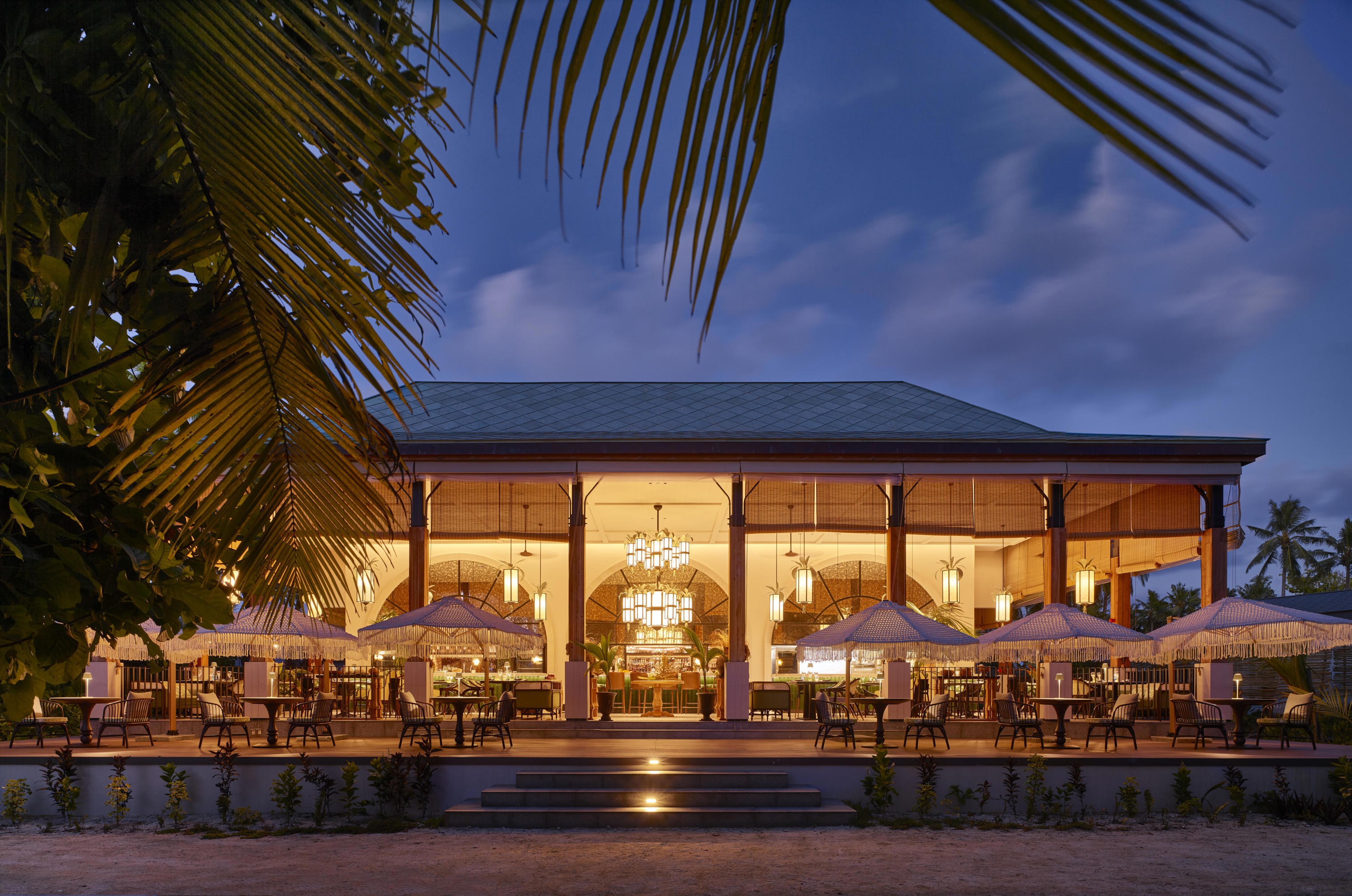 Exterior of Maison De Epices lit up at night with tables set out on the terrace framed by a palm tree
