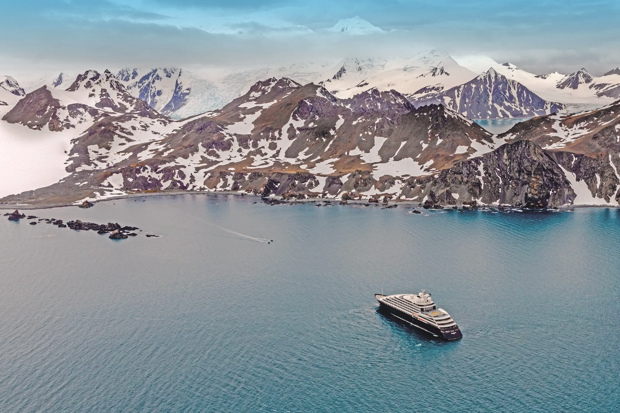 Luxury yacht anchored in a calm bay surrounded by snow-capped mountains and rugged terrain.