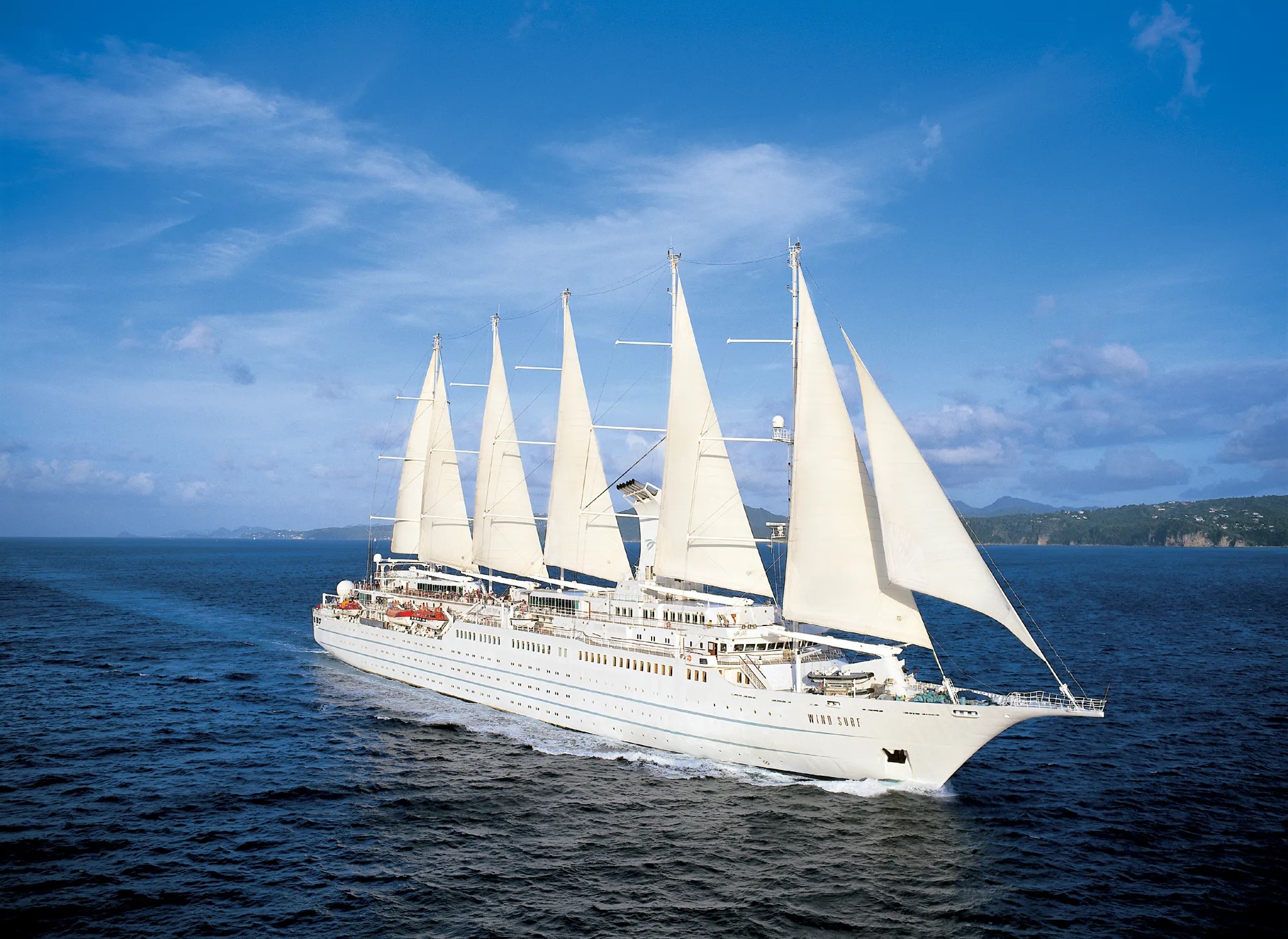 A large white sailing cruise ship with five tall masts and full sails gliding through the ocean under a clear blue sky.