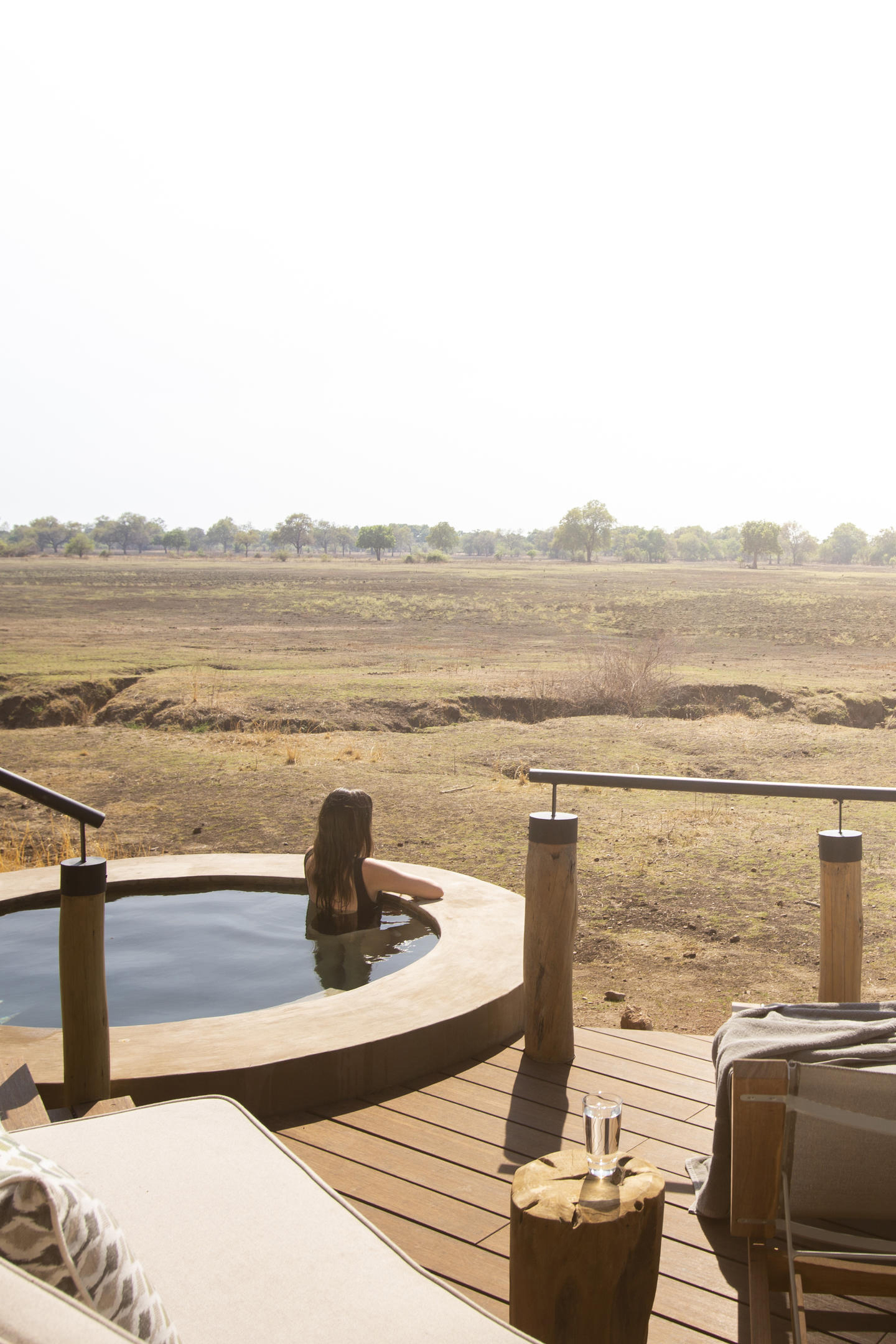 A serene view from Puku Ridge Camp showing a wooden deck with lounge chairs, a small round pool with a person sitting in it, and an expansive dry landscape stretching out to the horizon under a bright sky