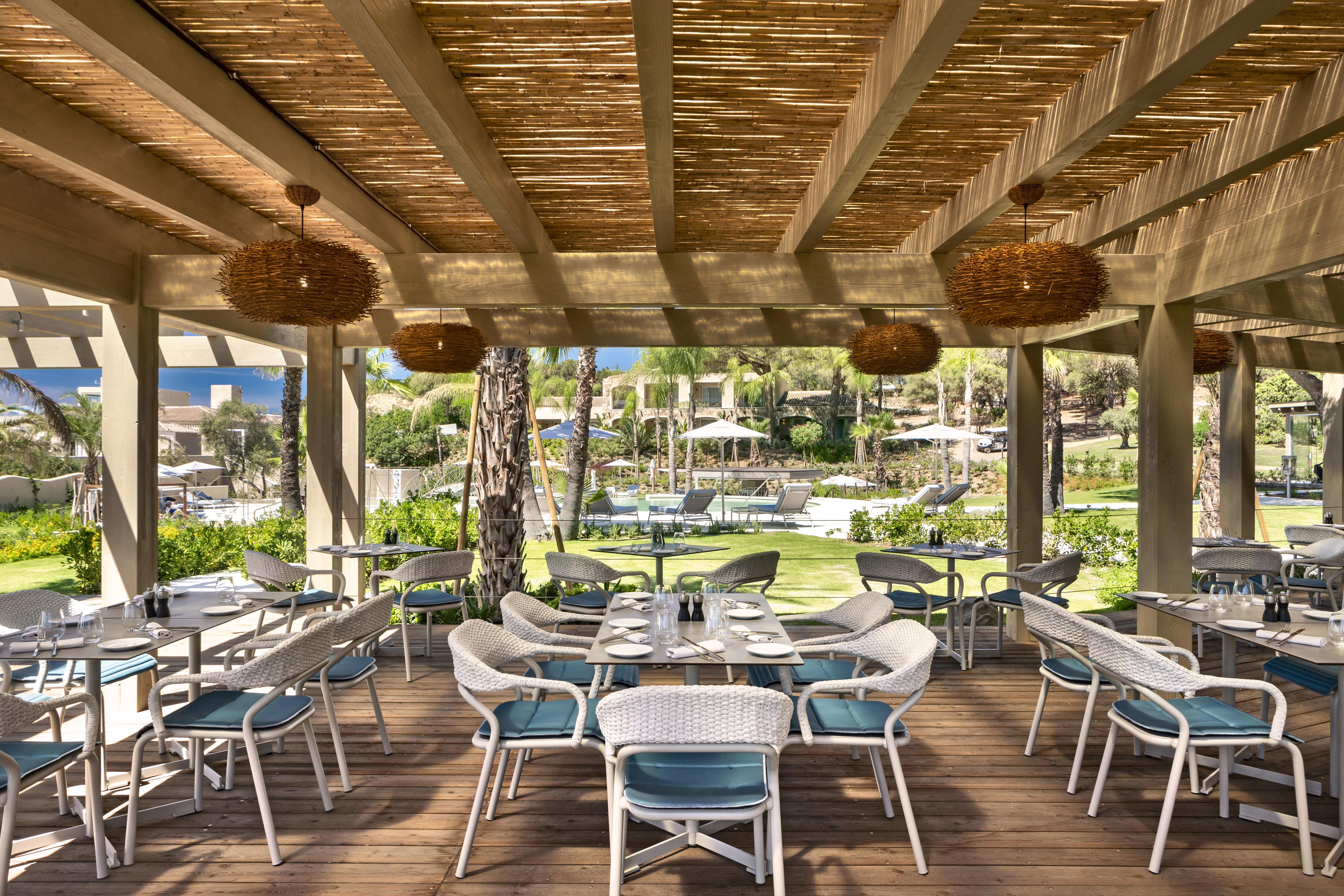 The interior of a restaurant below a thatched ceiling with woven light shades and white and blue tables