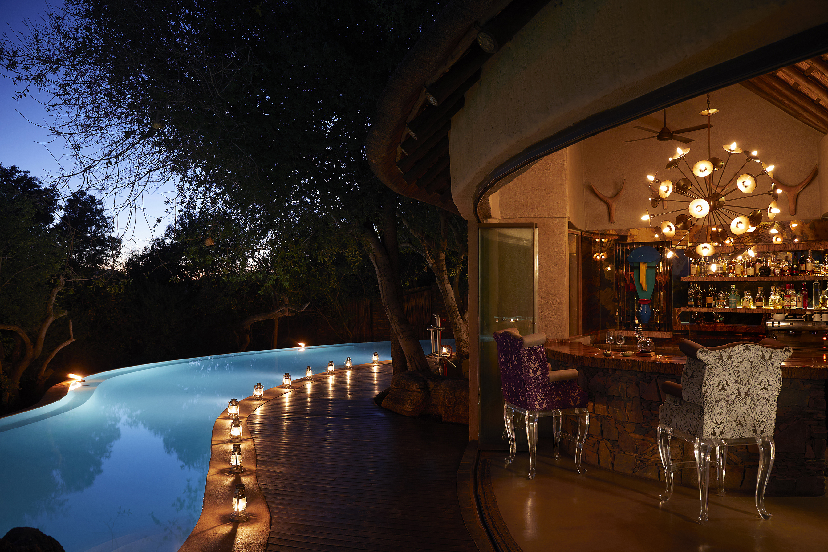 A decorative bar with stools and a statement light lit up at night beside a pool lined with candles