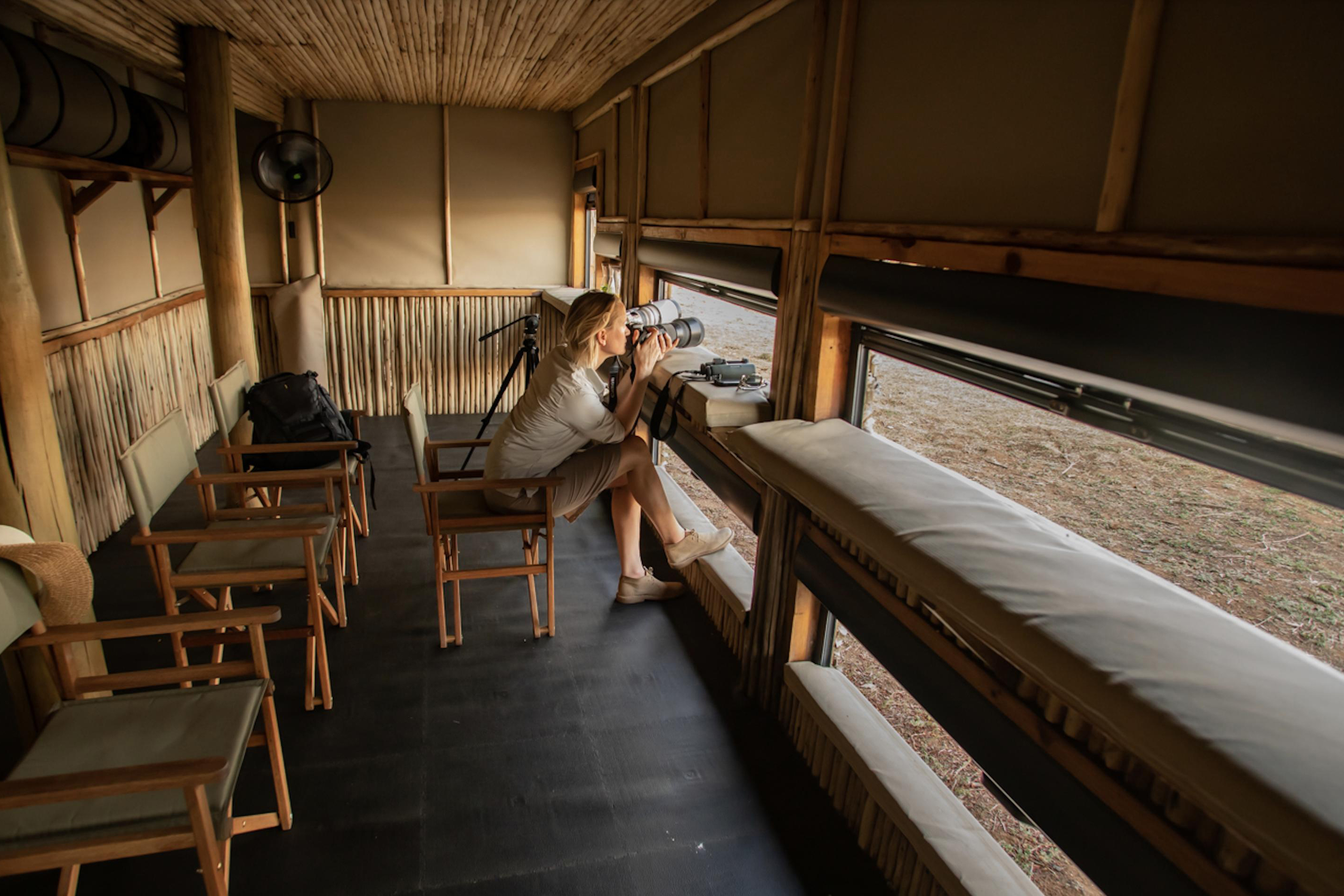 A person sitting inside a wooden observation hide at Puku Ridge Camp, using binoculars to view the landscape through an open window, with chairs and a backpack visible in the room
