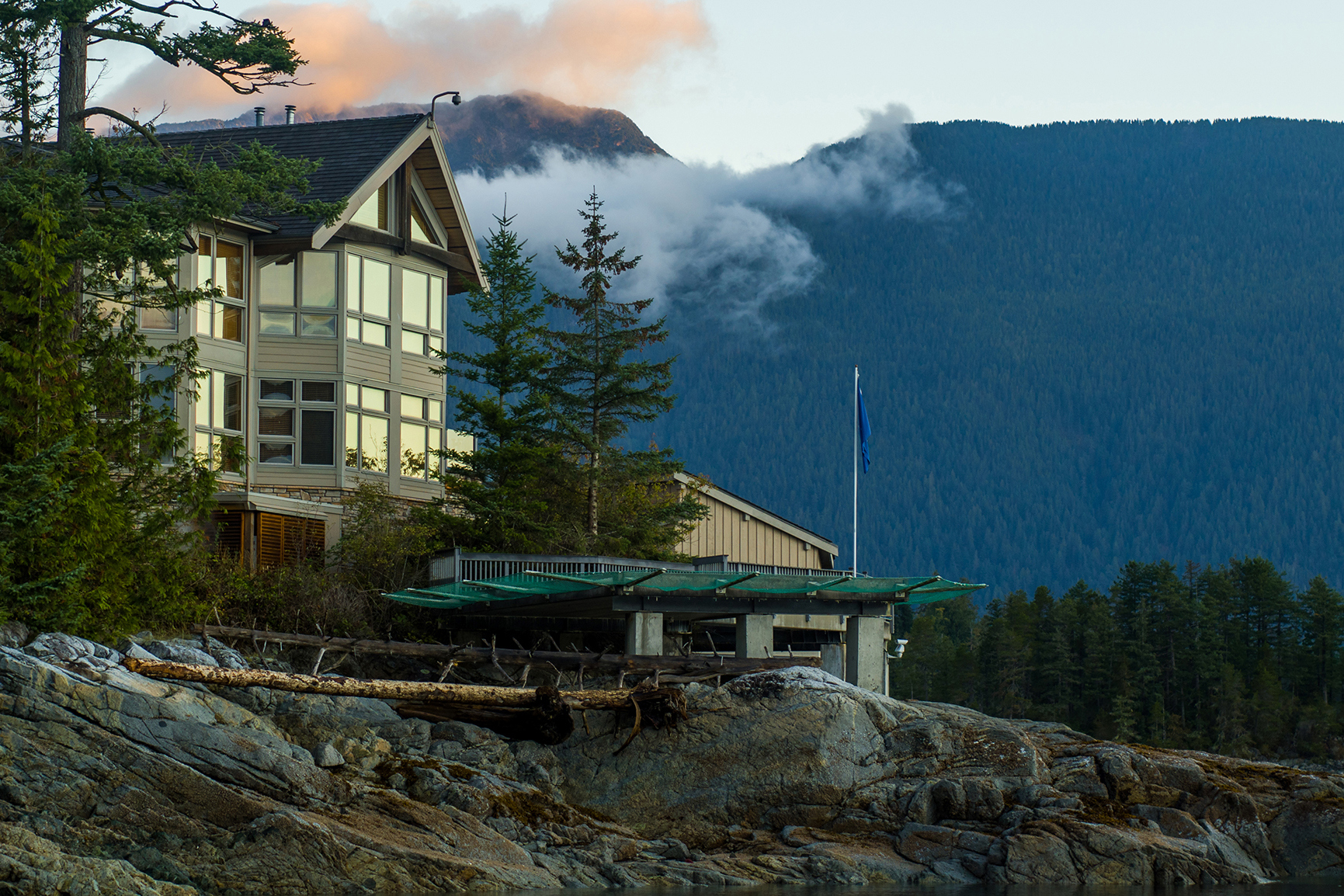 A wooden lodge perched on some rocks surrounded by trees with rocky mountains in the background