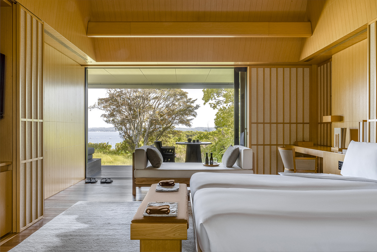 A pale wood panelled suite at Amanemu featuring a large bed, ottoman and day bed with a view to the terrace and forest beyond