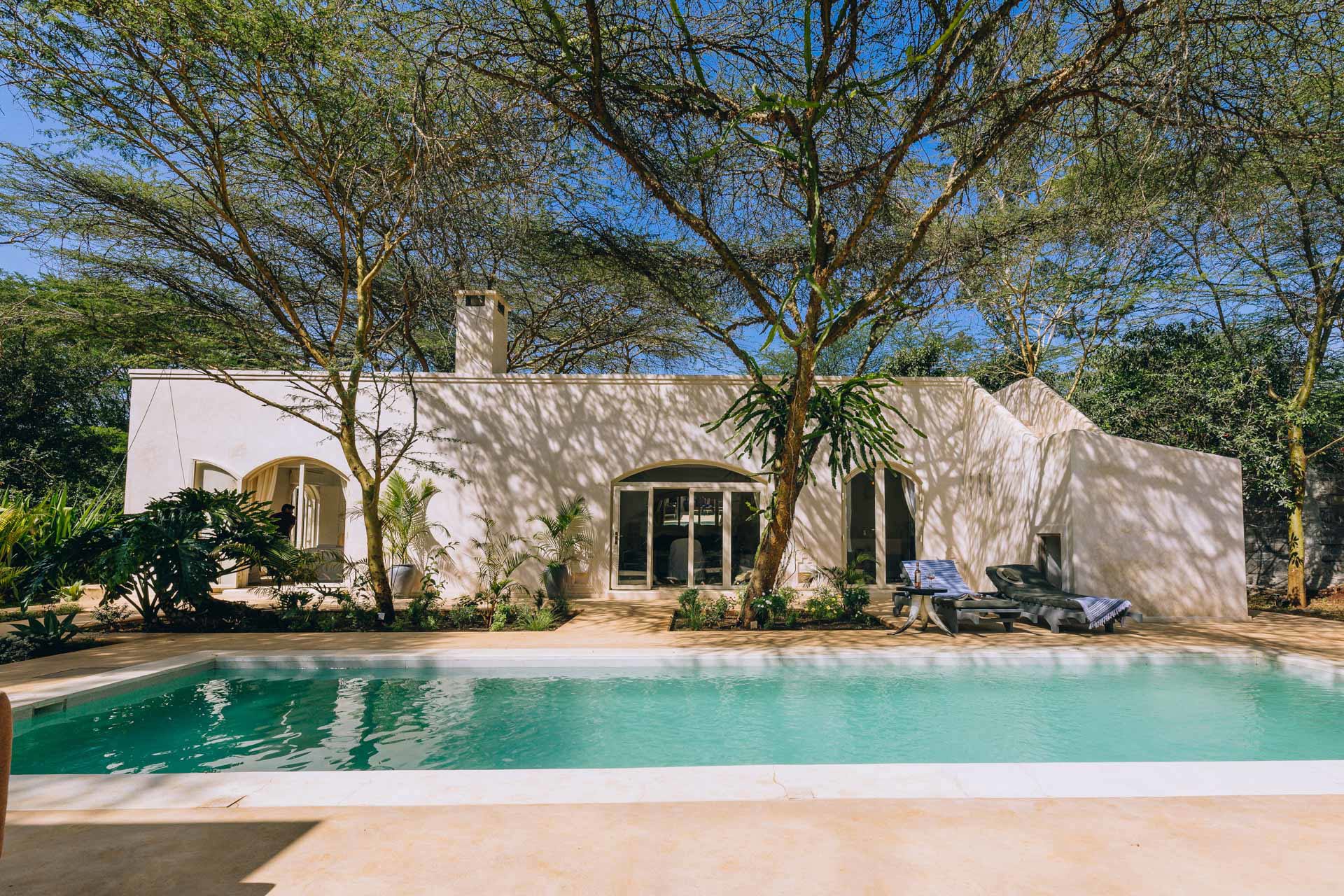 A whitewashed building at Manzili House shaded by trees with a pool and sun loungers in front