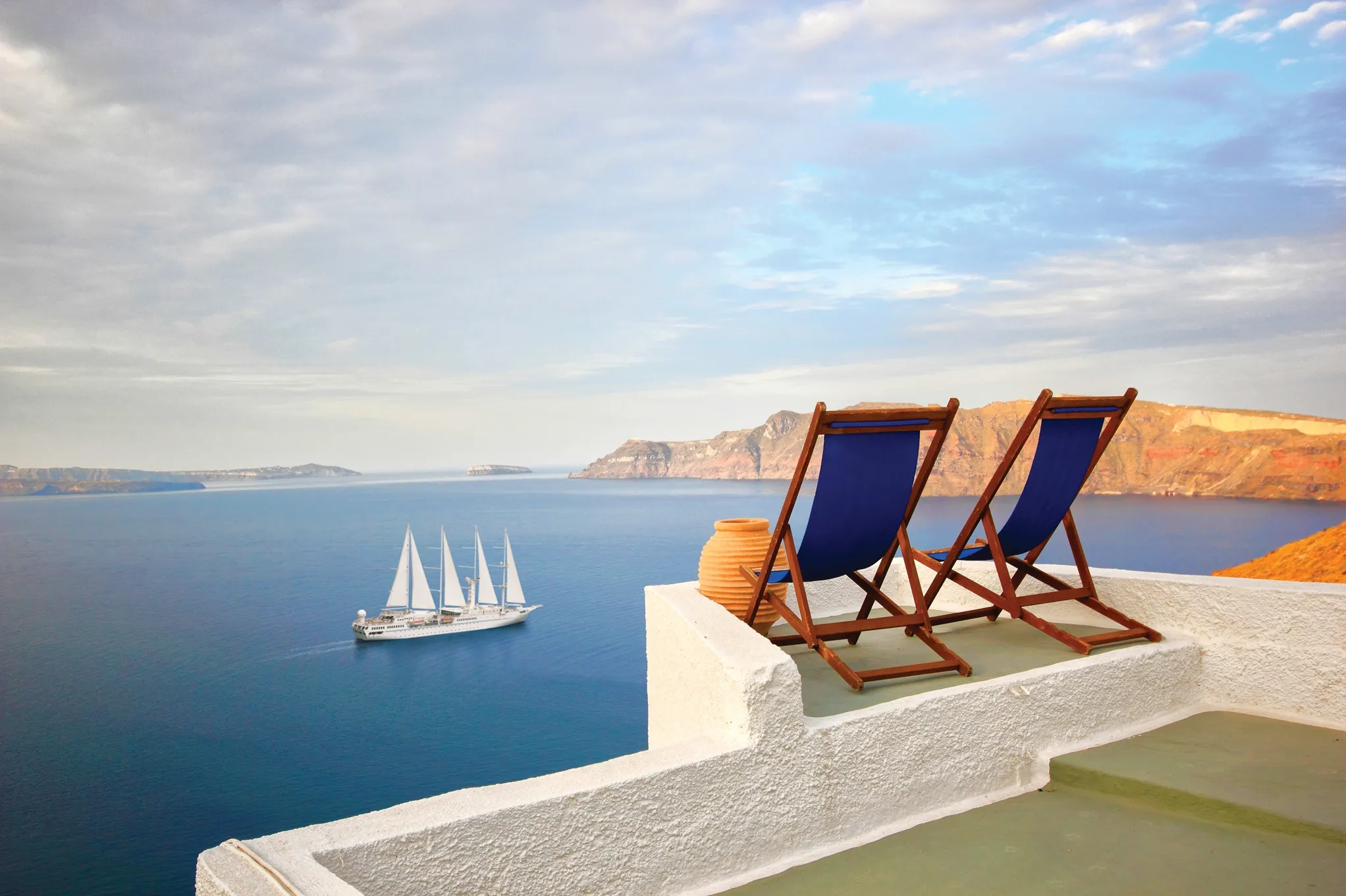 Two blue deck chairs and a clay pot on a white terrace overlooking the sea with a sailing ship and distant cliffs under a partly cloudy sky.