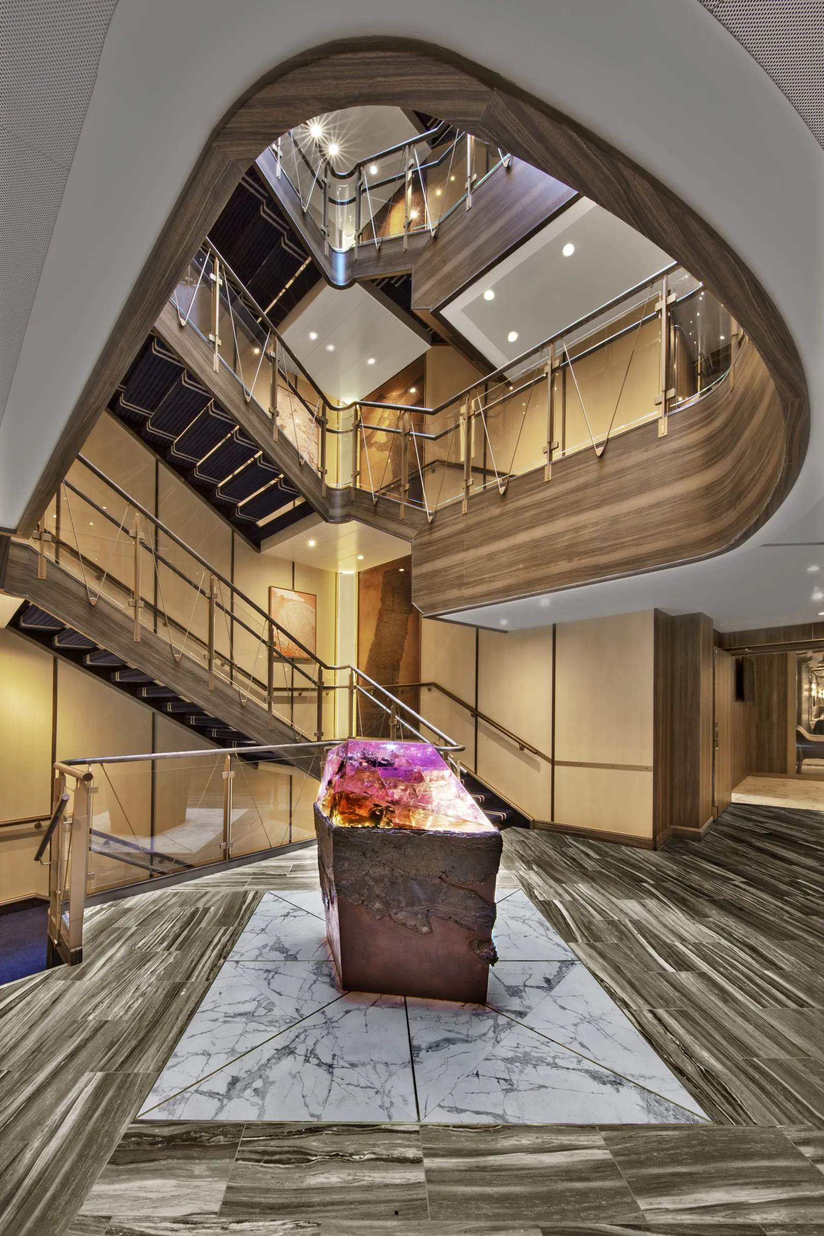 Modern staircase with wooden accents and glass railings encircling a colorful crystal sculpture on a marble floor.