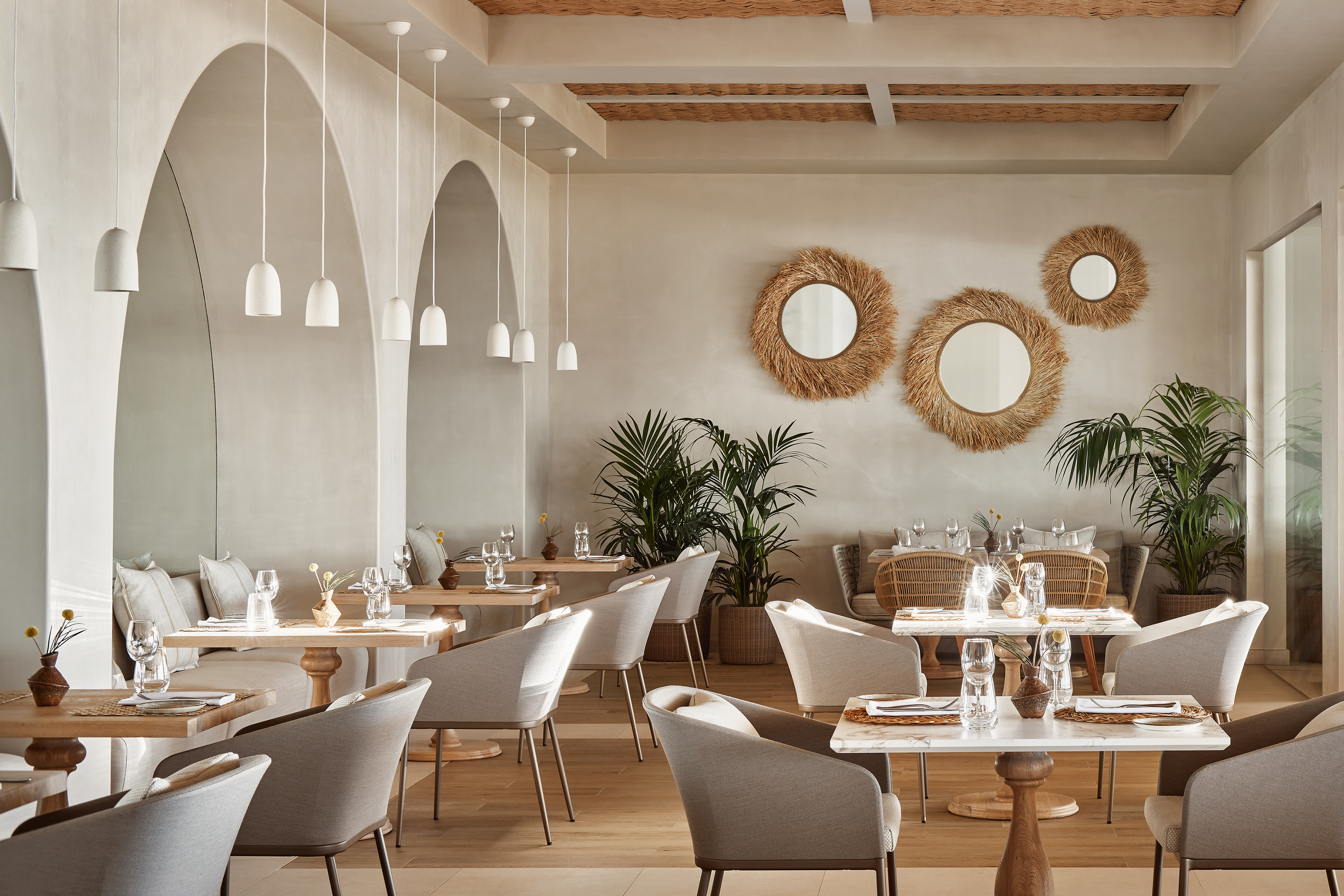 Interior of Fresco restaurant at Ikos Olivia featuring plasterwork walls with circular straw mirrors and comfy chairs and tables set for dinner