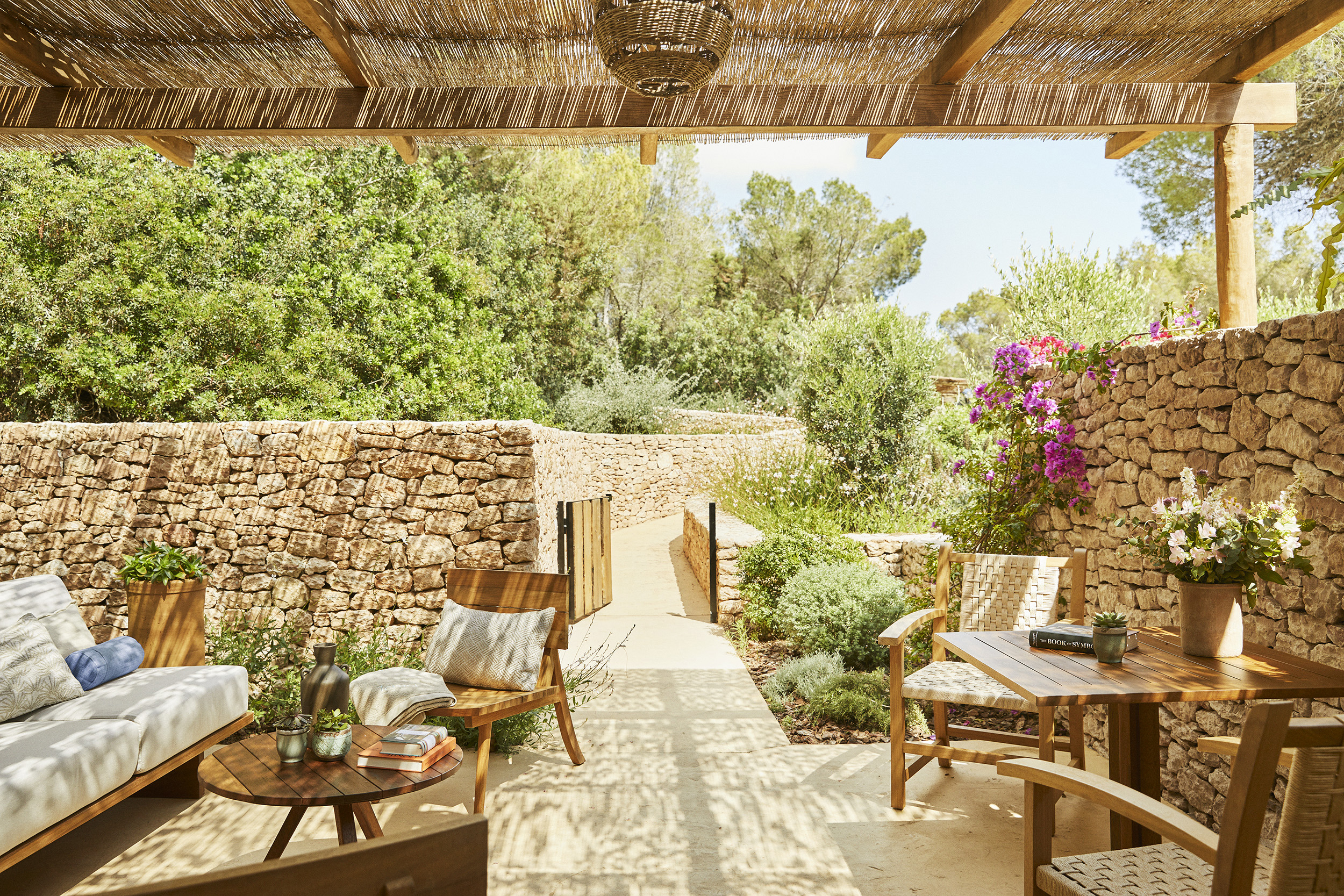 The terrace of a double deluxe room at Teranka with wooden furniture decorated with cream cushions and tables decorated with books and vases beneath a woven canopy