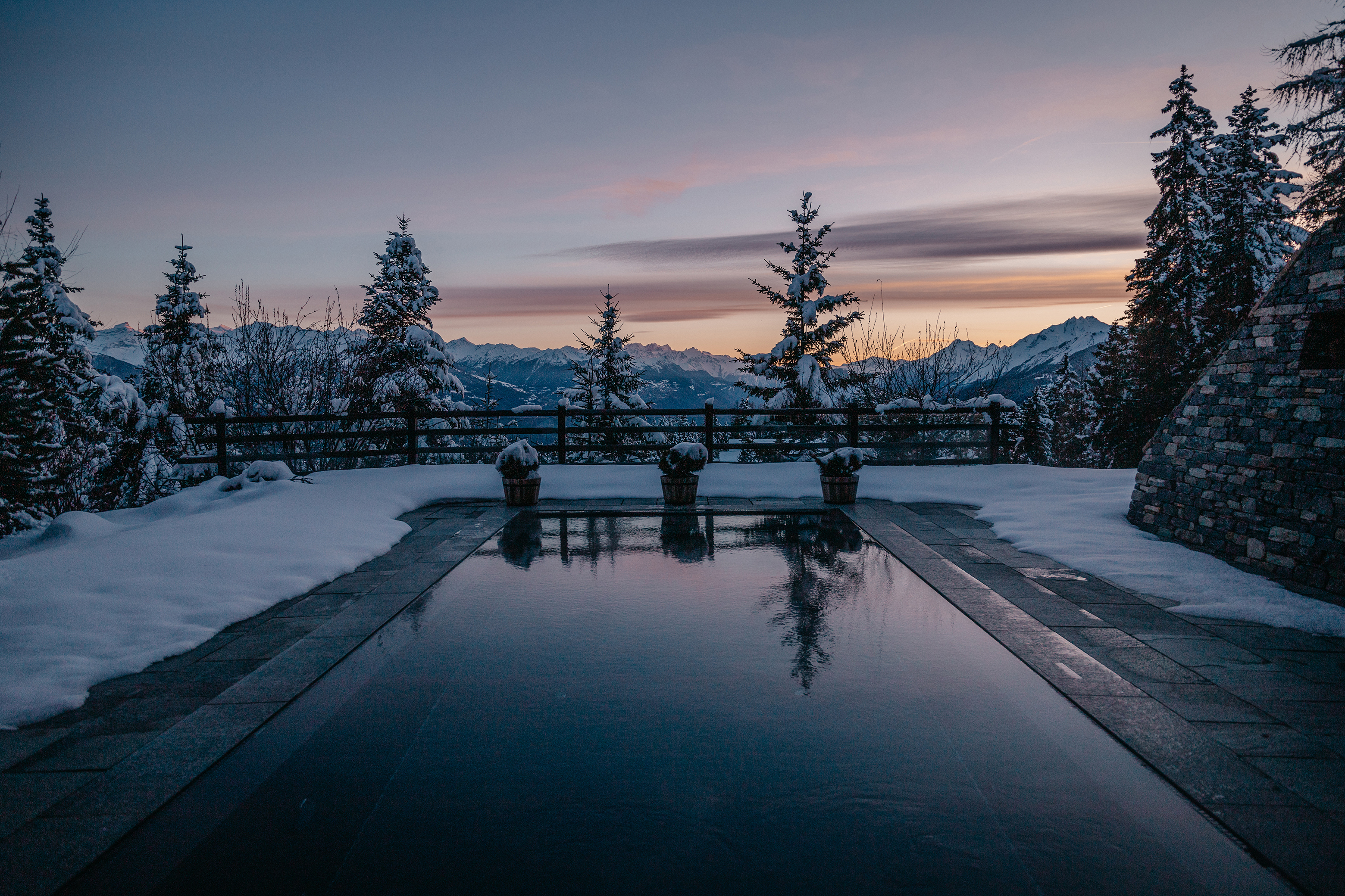 A snow-covered outdoor pool terrace surrounded by trees at sunset