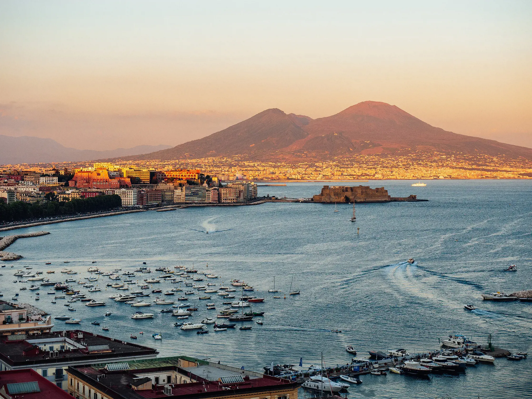 Panoramic view of Naples waterfront with boats in the bay and Mount Vesuvius in the background at sunset.