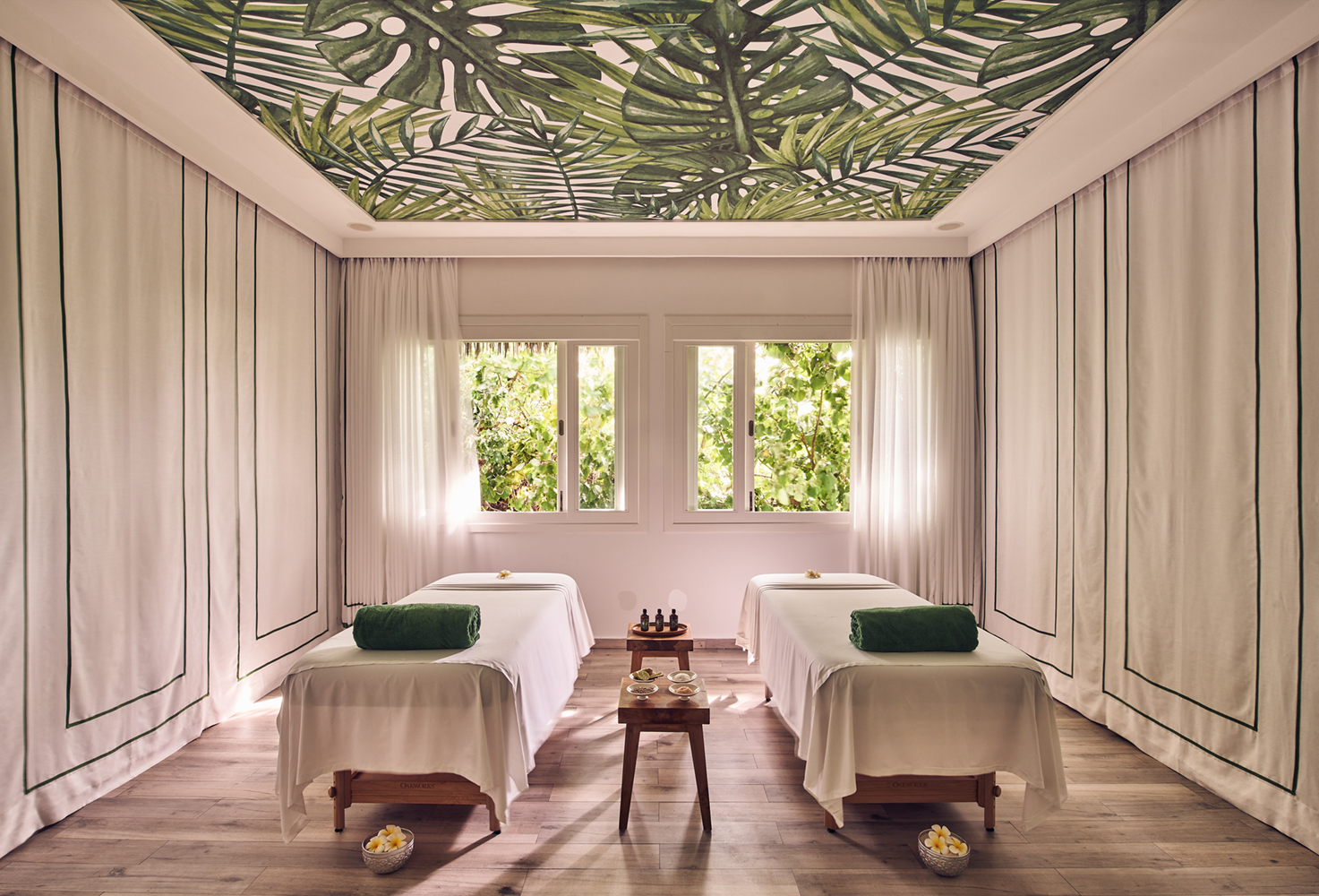 The airy treatment room of the spa at Finolhu featuring two massage beds with white and green towels in the centre of a room with white fabric walls and leaf patterned ceiling with windows ahead