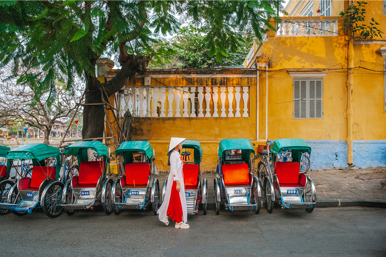 A person in a white robe and conical hat walking past a row of colourful rickshaws by a yellow building and green tree.